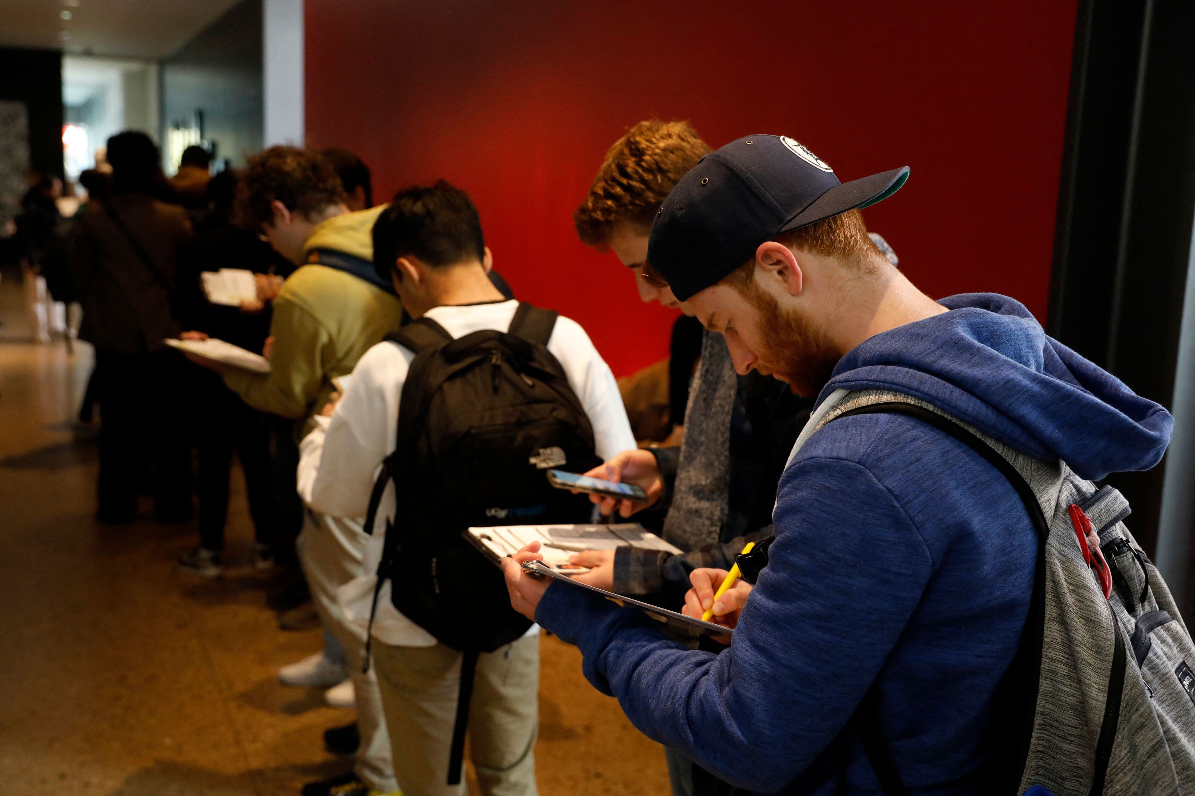A man in a sweatshirt and backpack stands in line and write on a clipboard