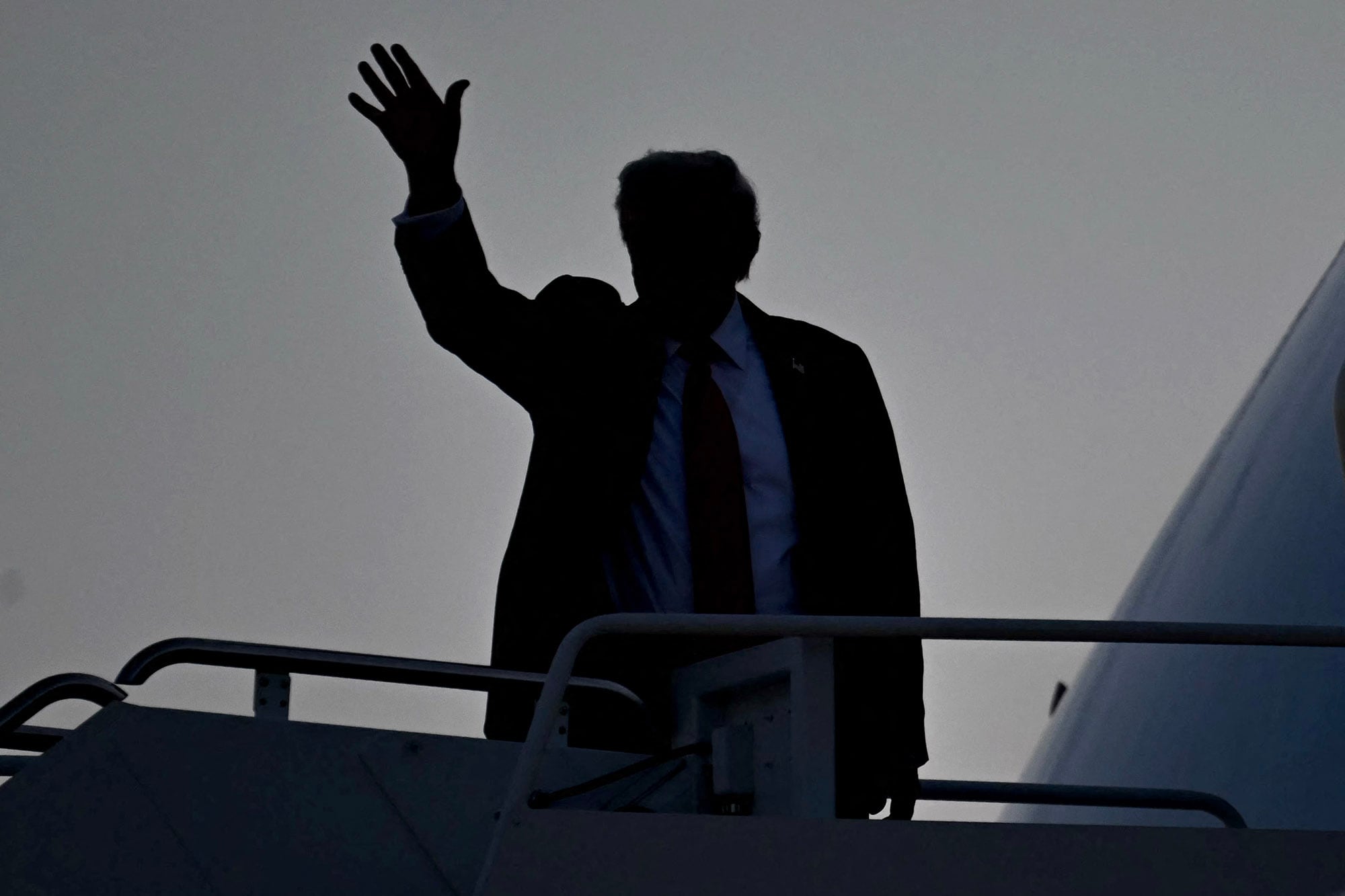 A photograph of a silhouette of President Donald Trump waving while standing outside on an airplane ramp.