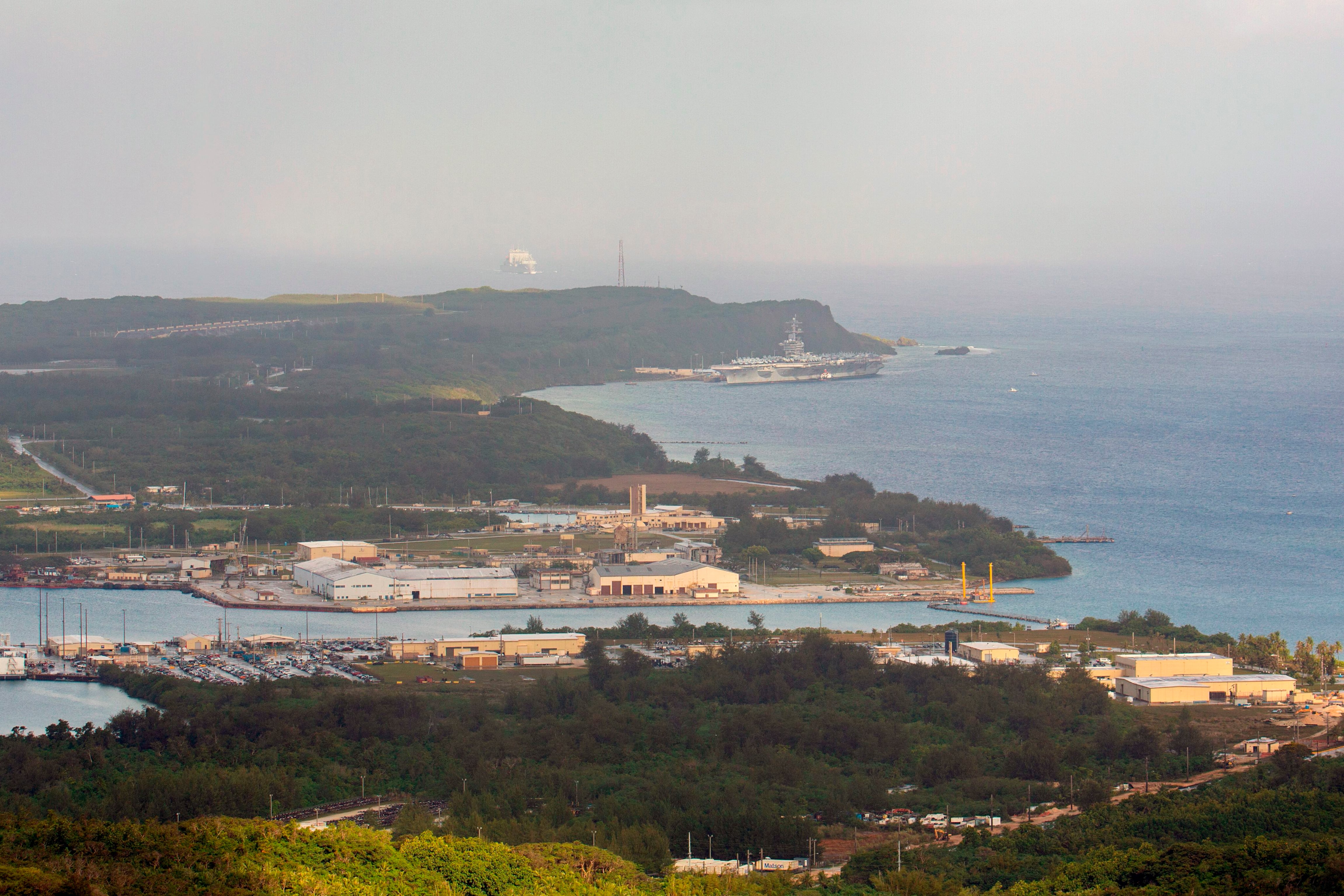 A far away view of green land and the ocean with various buildings and U.S. military ships and buildings.
