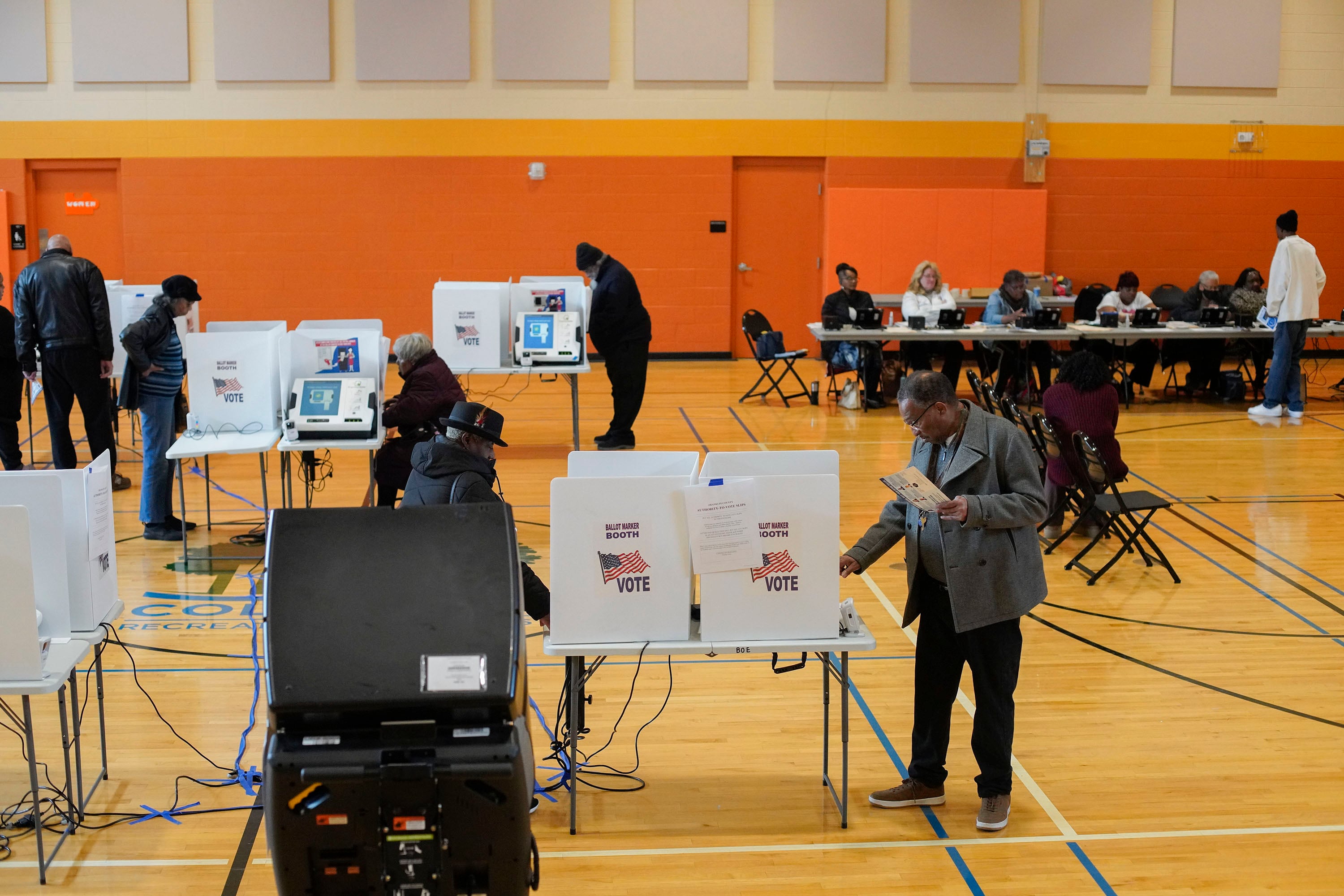 In a large gymnasium with an orange wall in the background, several people stand near voting booths in the middle and front of the frame with several people sitting in a row behind a table.