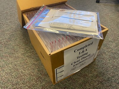 A cardboard box full of election testing documents sits on the floor with carpet.