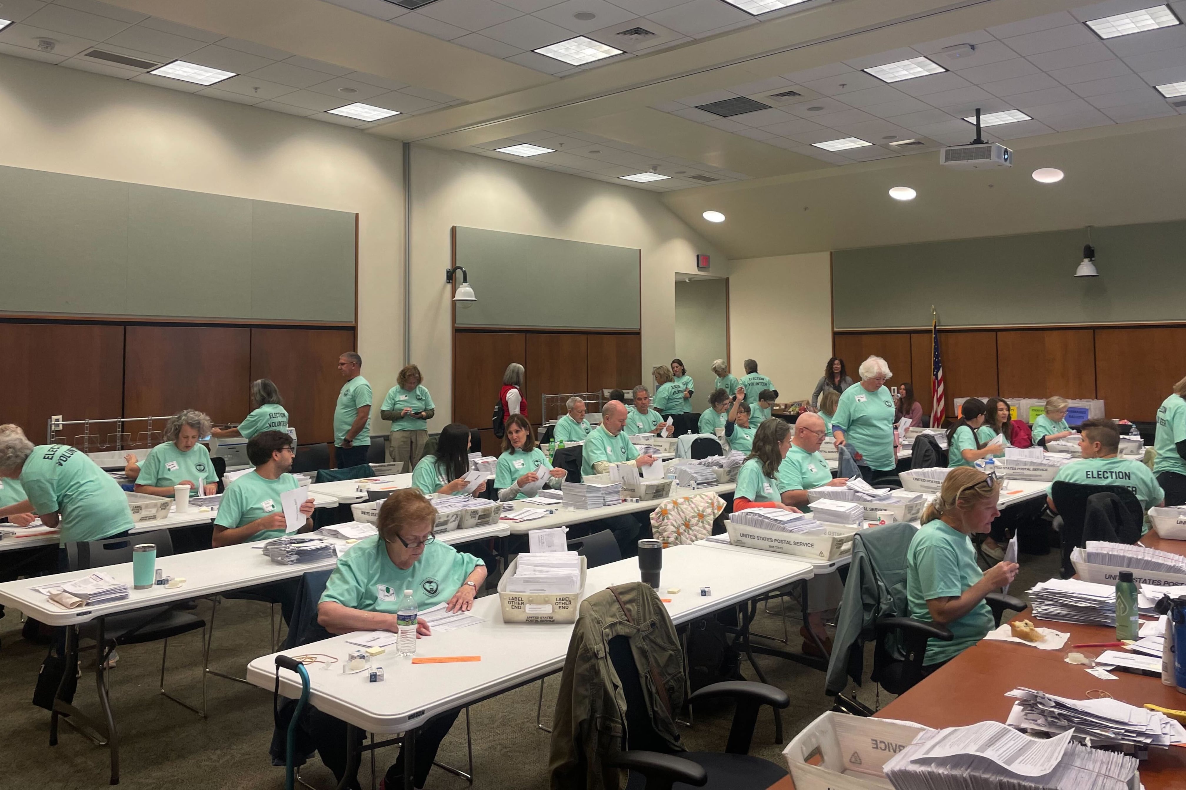 Workers in green shirts work at long tables covered with ballots and bins 