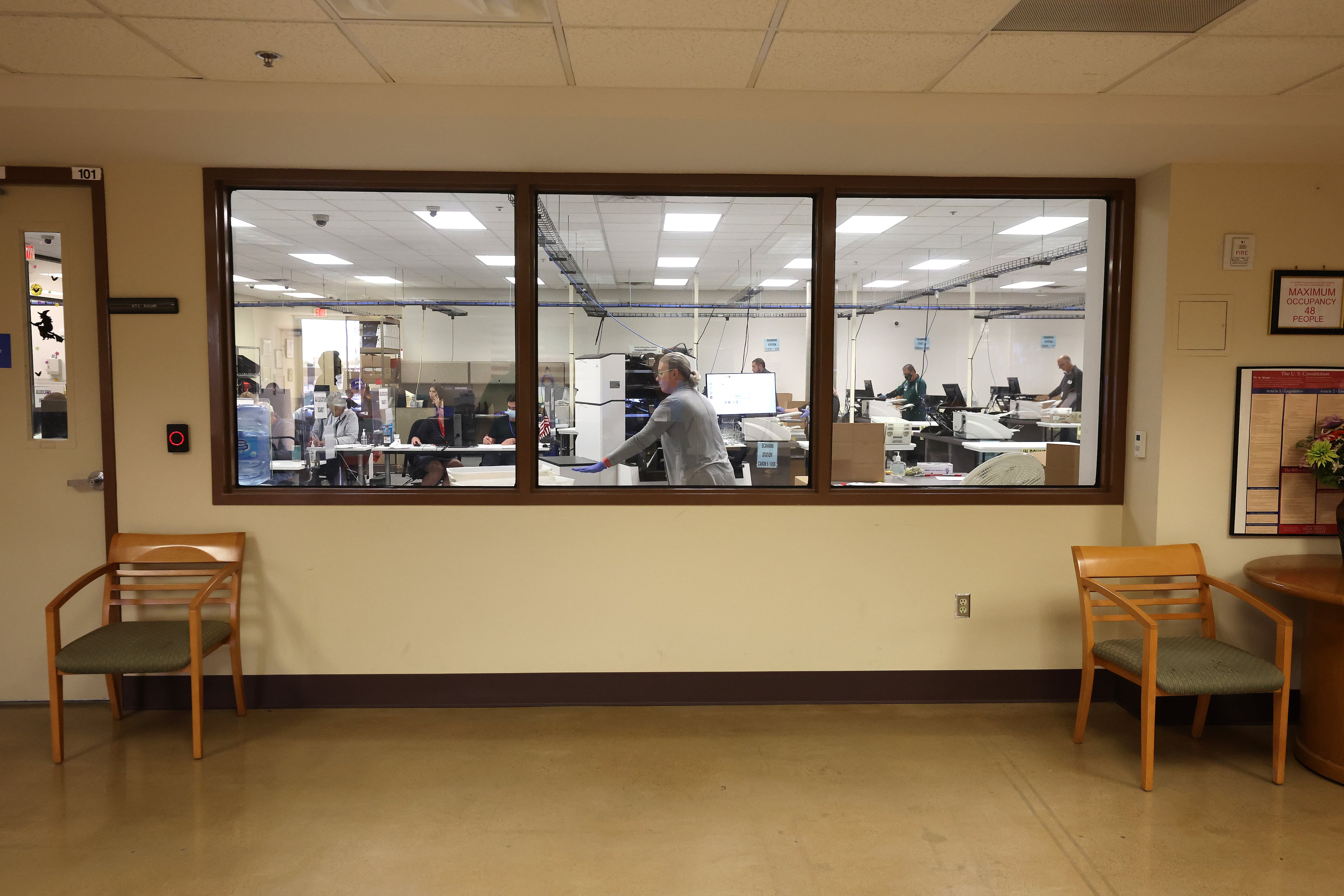 A woman with glasses wearing a gray sweater standing behind a glass window feeds paper into a machine.