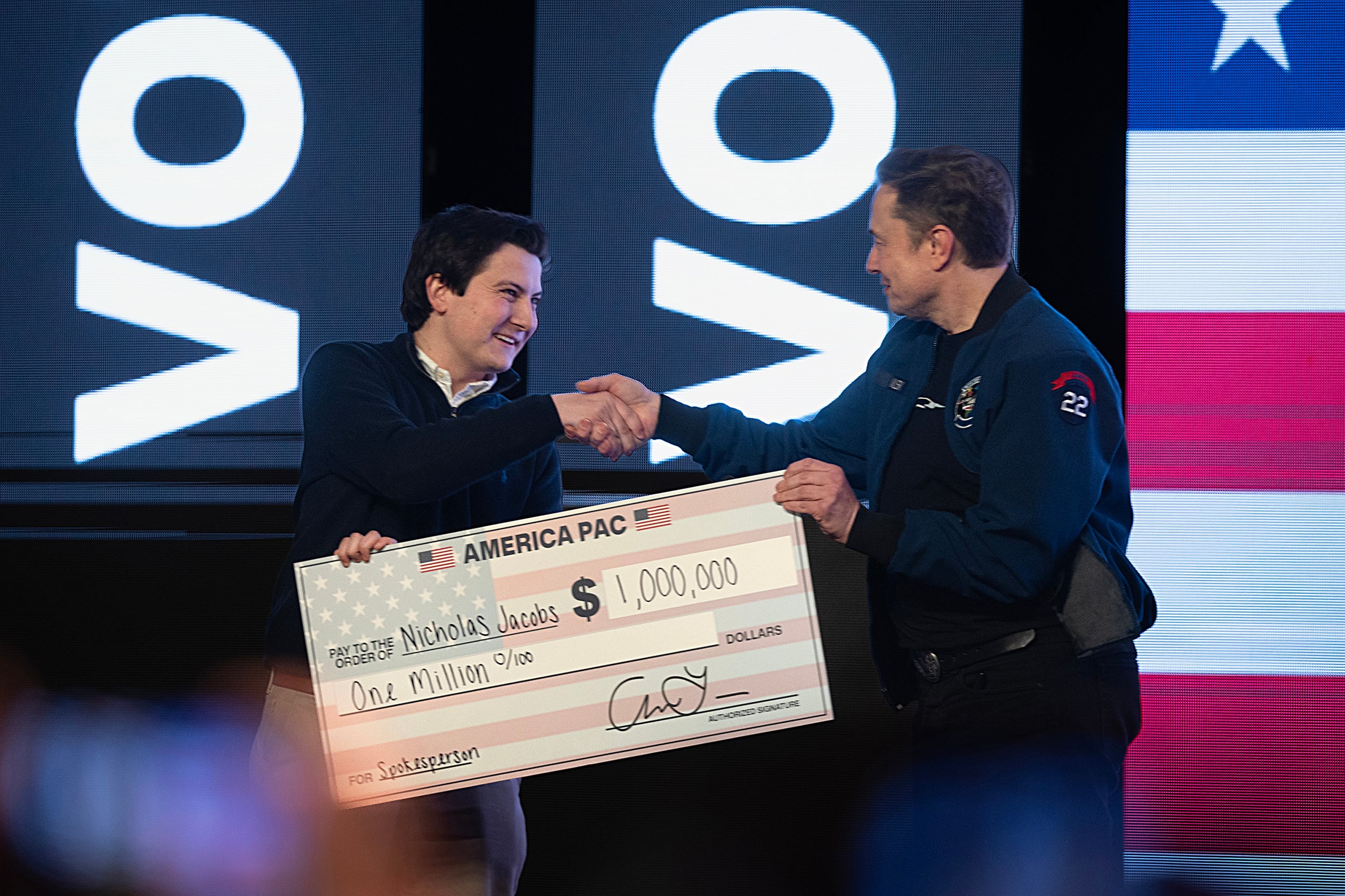 Two white men shake hands on stage and exchange a check of one million dollars.