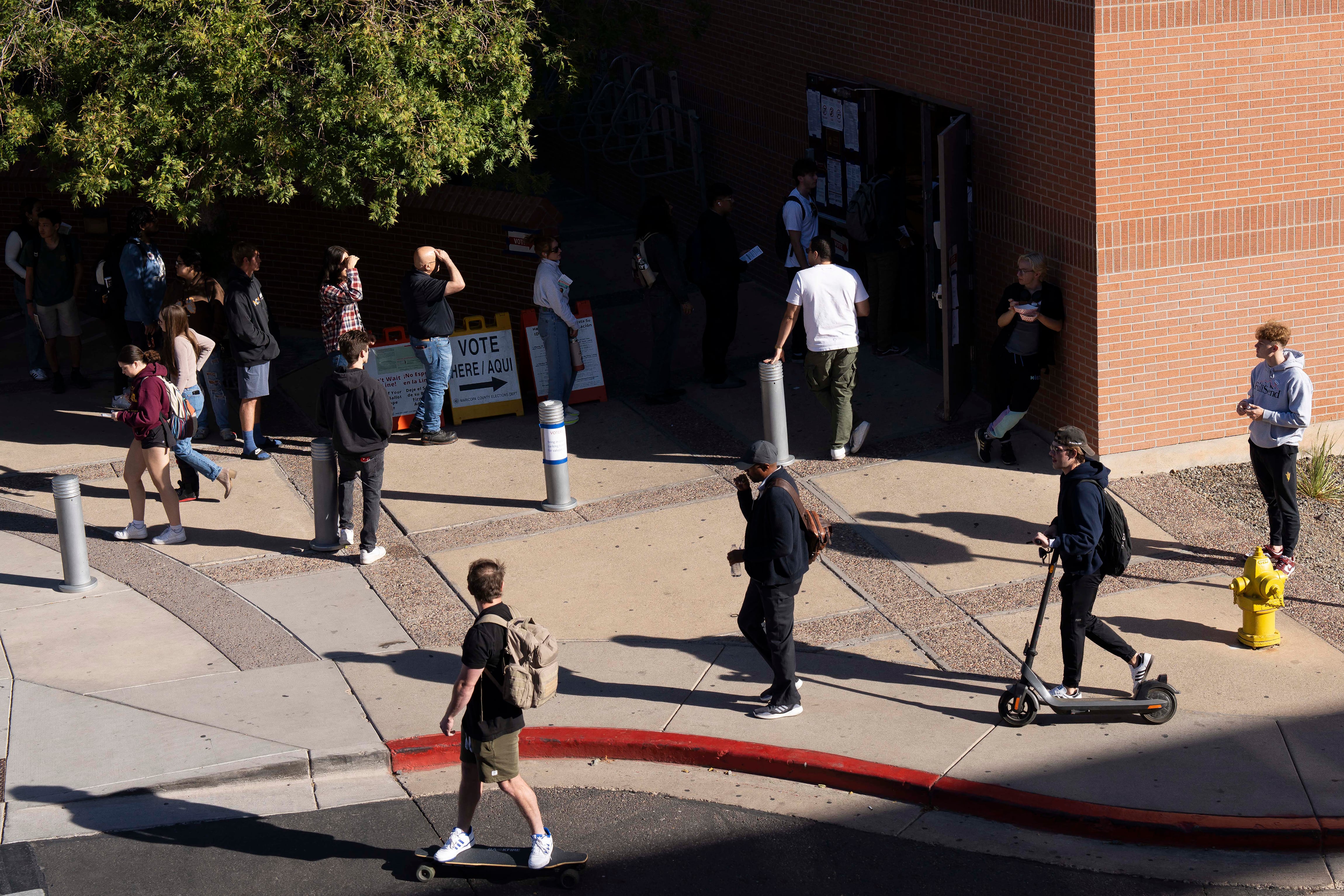 A group of college students walk by other student voters standing in line outside of a brick building on a sunny day.