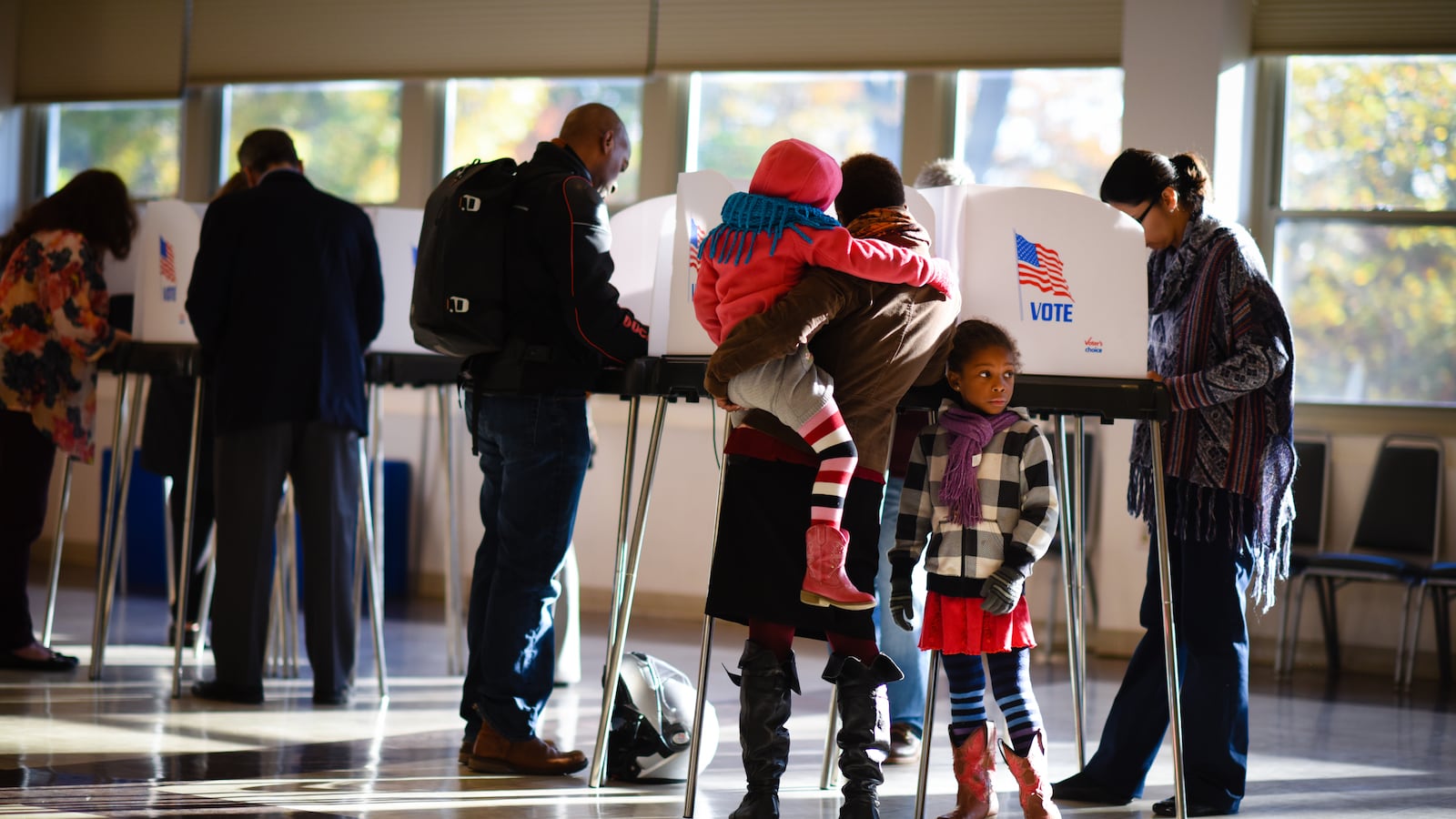 A woman holding a small child stands at a row of polling place booths, along with other voters, as a girl looks on.