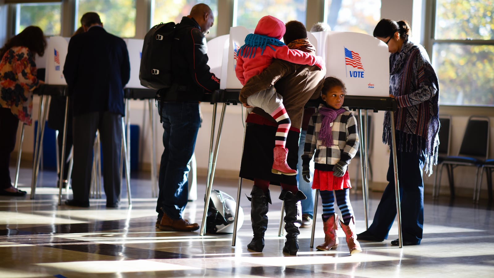 A woman holding a small child stands at a row of polling place booths, along with other voters, as a girl looks on.
