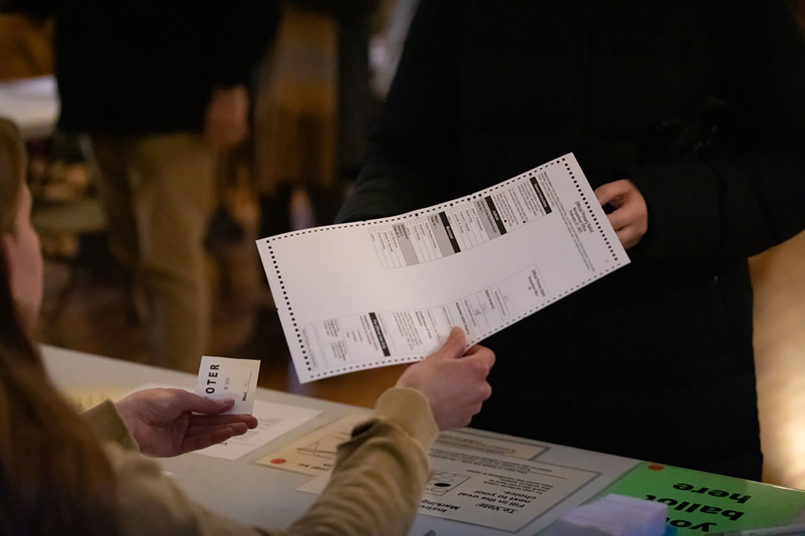 A close up of a person handing a ballot to another person at a table.