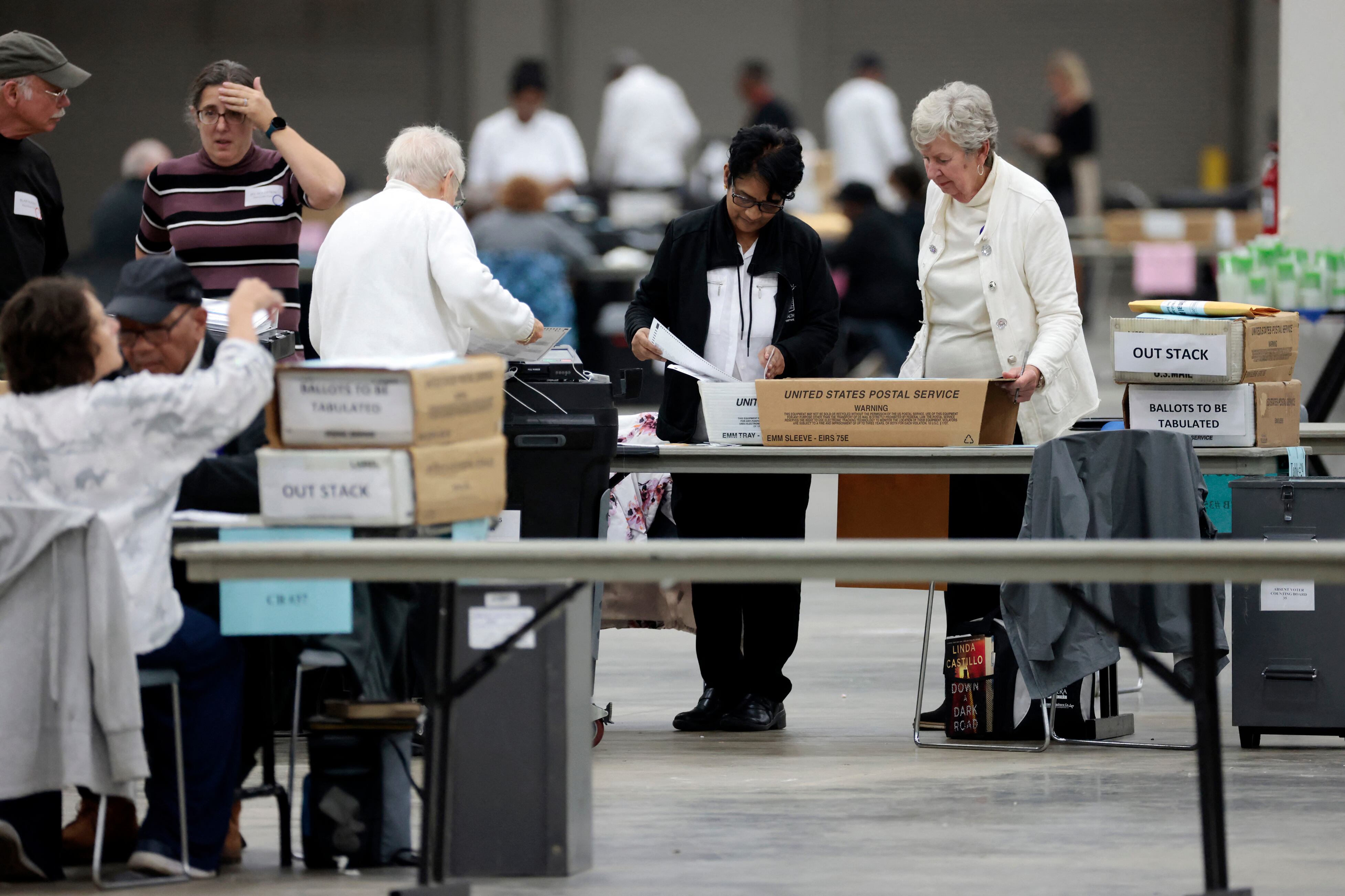 A group of adults stand near boxes of ballots and tables in a large room.