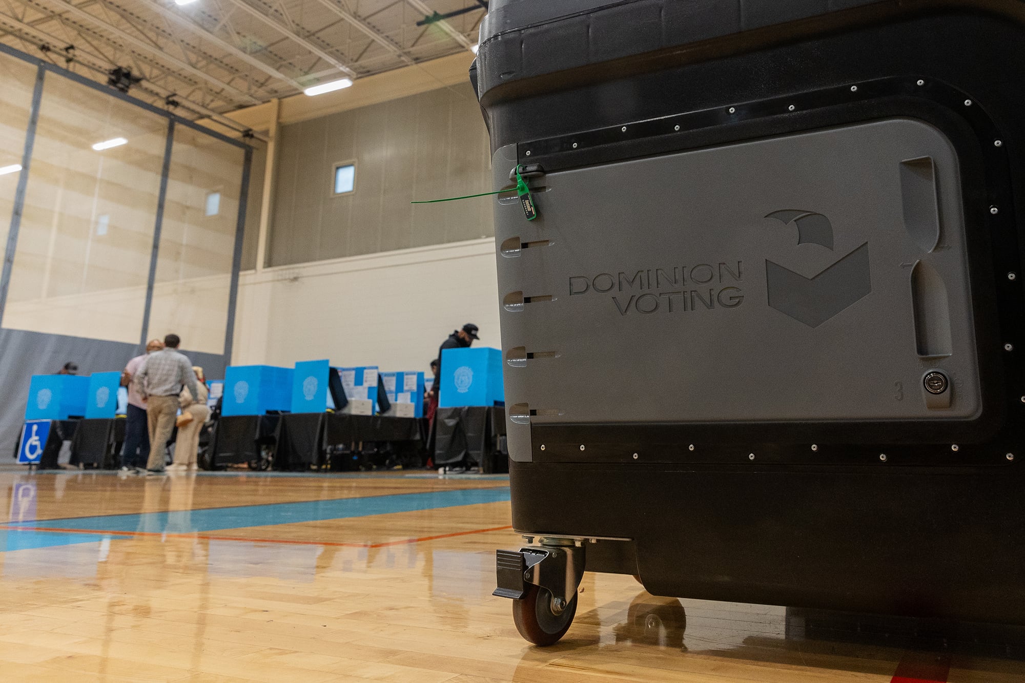 A photograph of a black and grey box on wheels in a large gym-like room with people voting in the background.