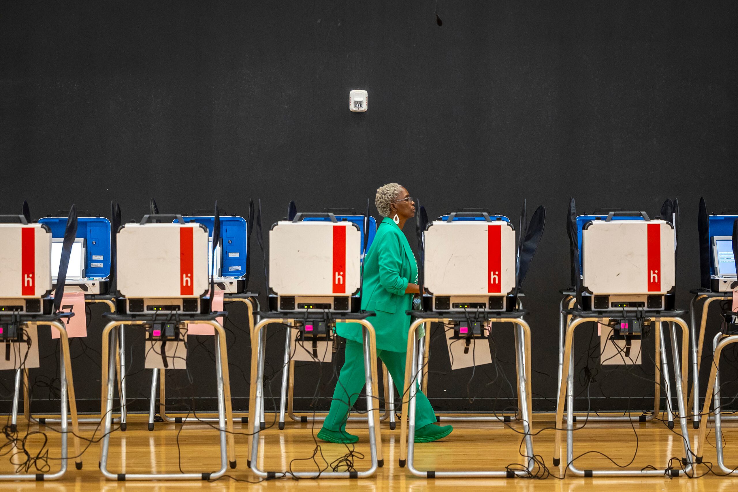 A photograph of a Black woman walking next to a row of voting machines.
