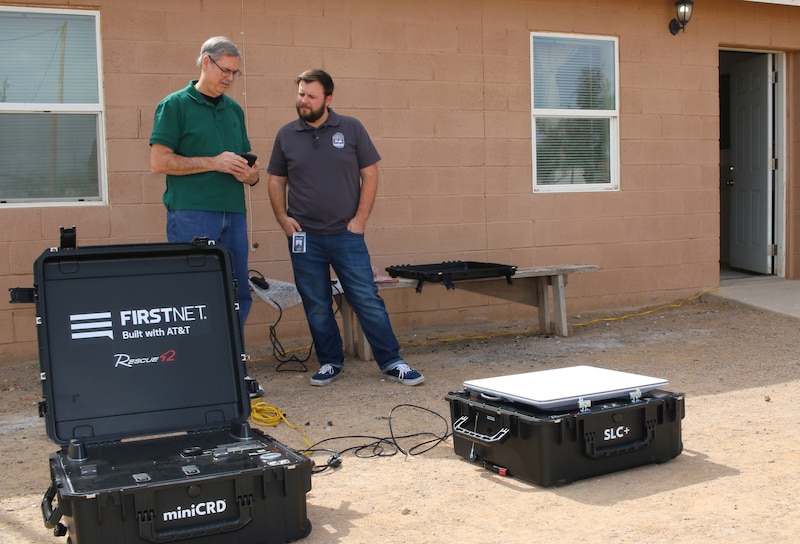 Two people stand next to each other looking at a cellphone with two large black boxes in the foreground and a tan house in the background.