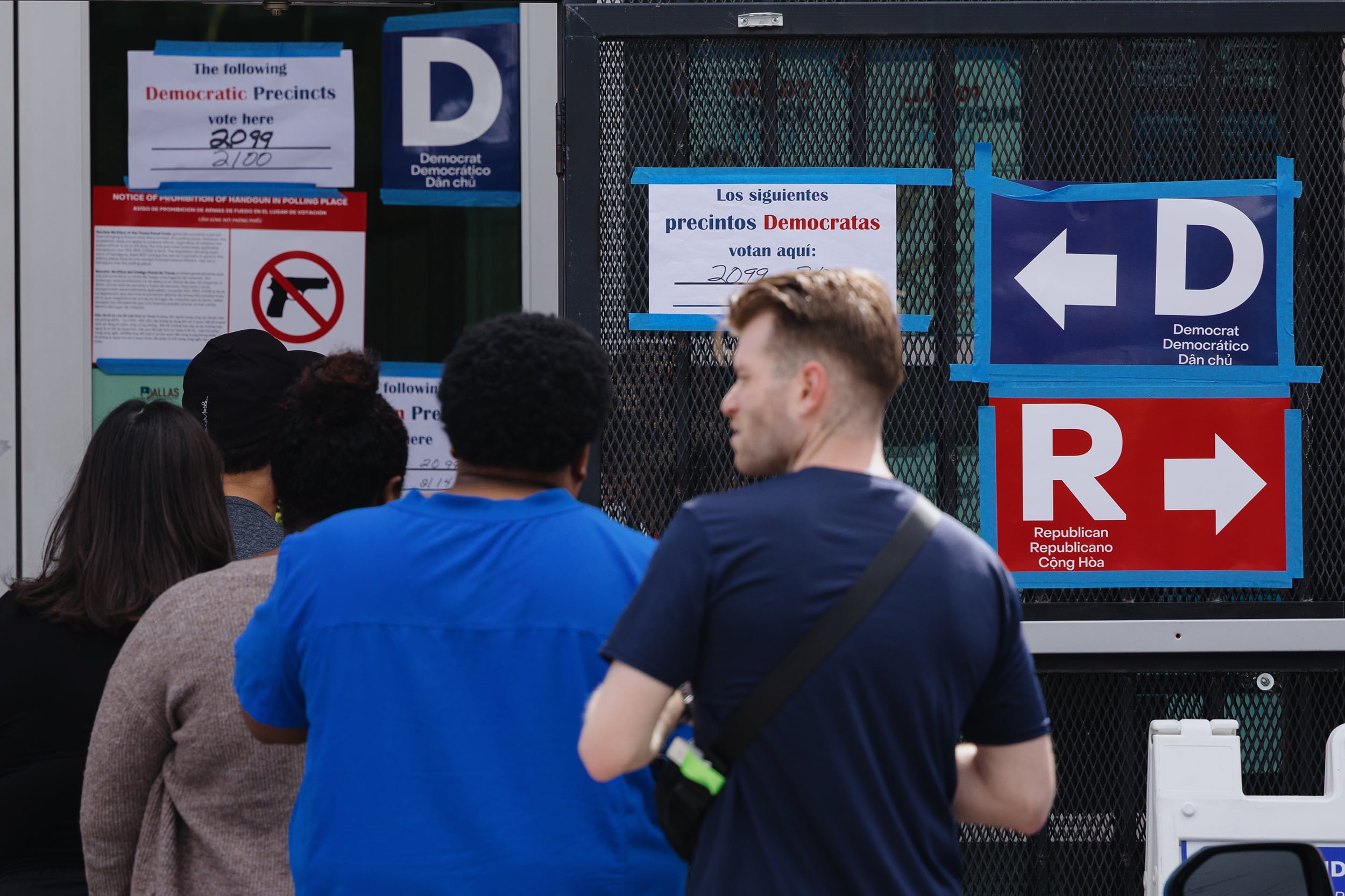 A photograph of a line of people standing in line facing a building with glass doors and different signs on the side.