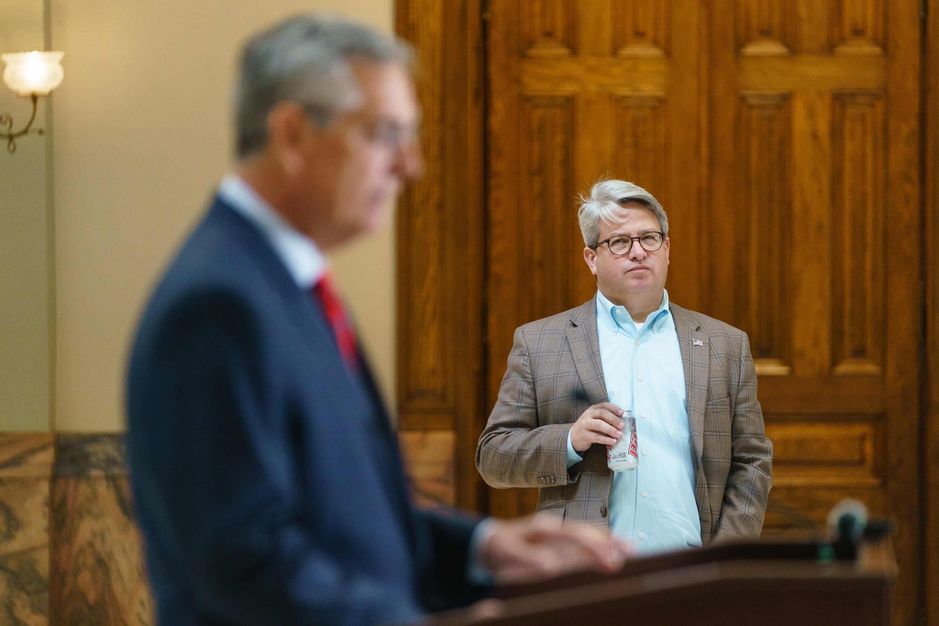 Two men wearing suits, one in the foreground speaking from a podium and one stands in focus in the background holding a carbonated drink.
