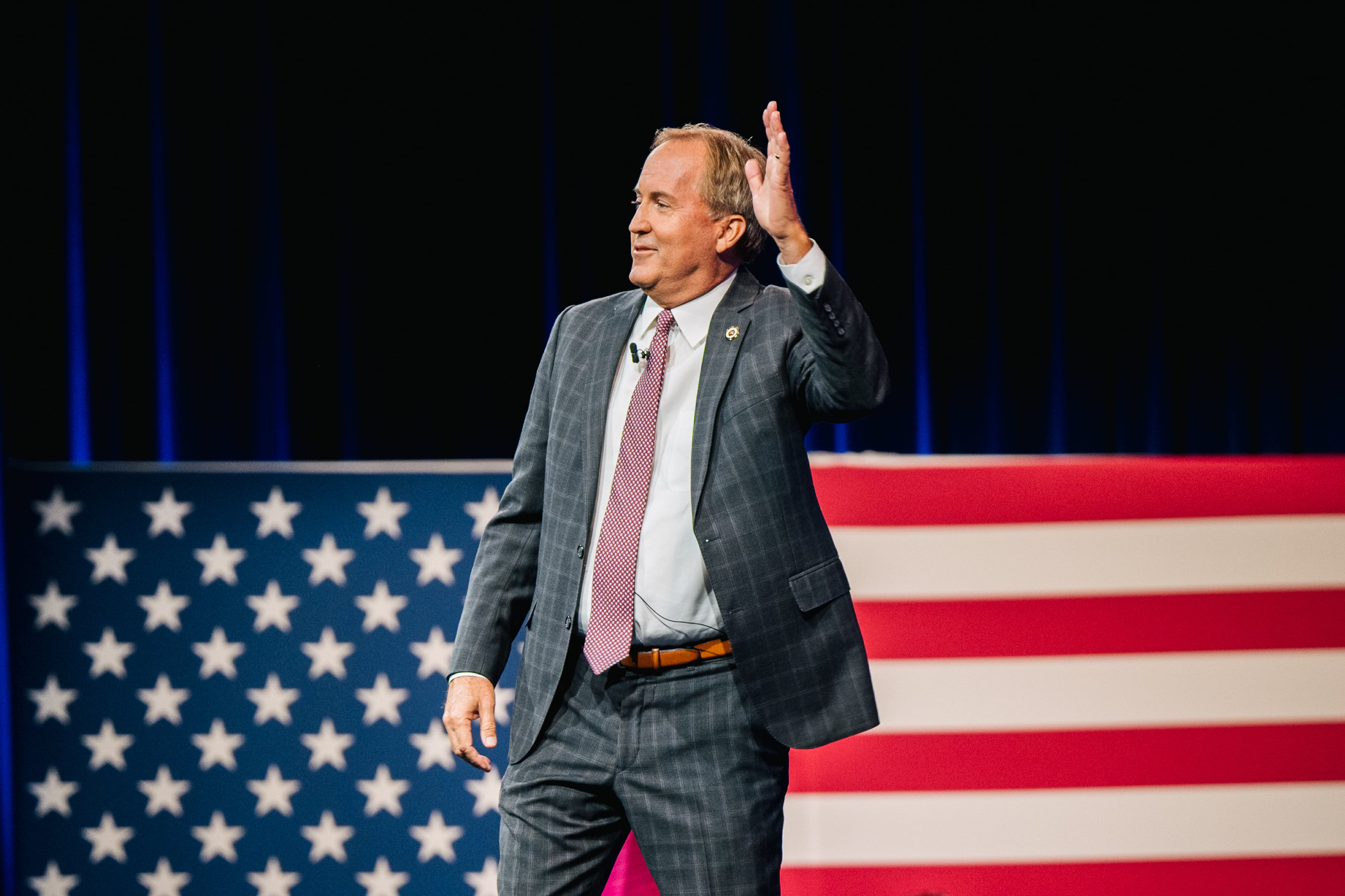A man in a suit and tie waves his hand against the backdrop of an American flag.