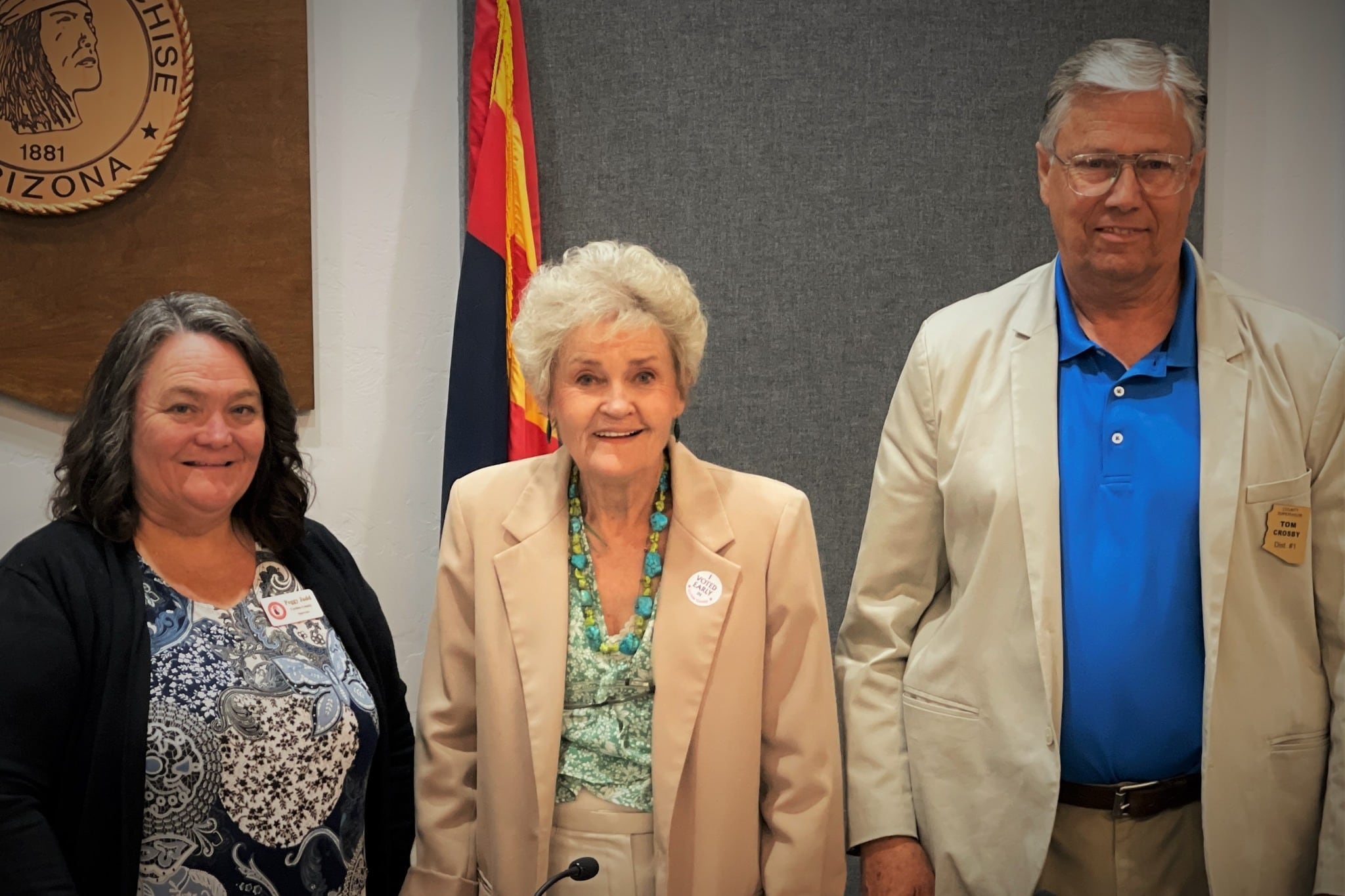 Three people with coat jackets stand and look at camera with a flag and emblem in the background.