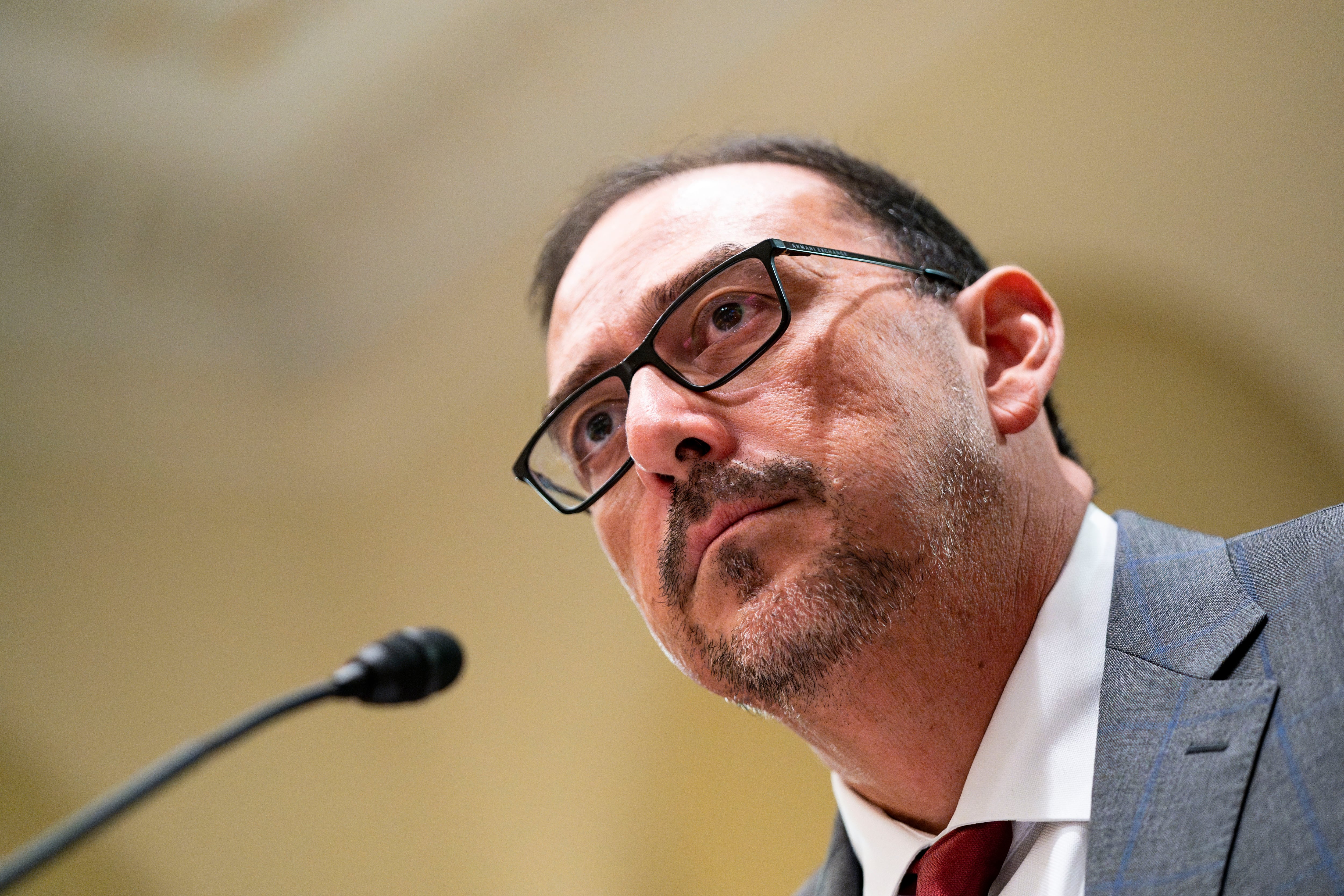 A seated man in a suit and glasses looks into the distance while a small microphone nears his face.