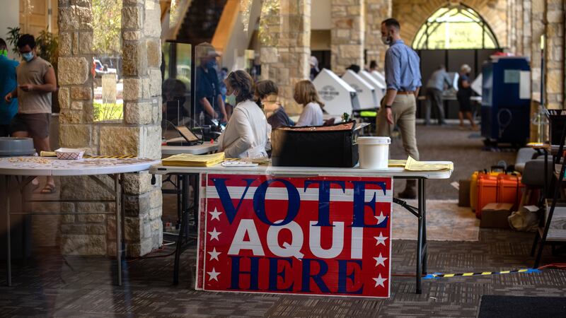 A sign reading “vote here aqui” hangs from a table in front of a row of polling stations and poll workers