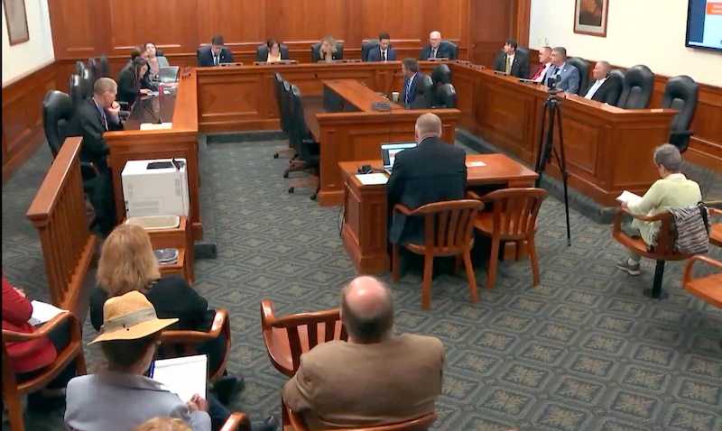 A man in the middle of a conference room sits at a wooden table while a large group of people sit around the edge of the room looking at him.
