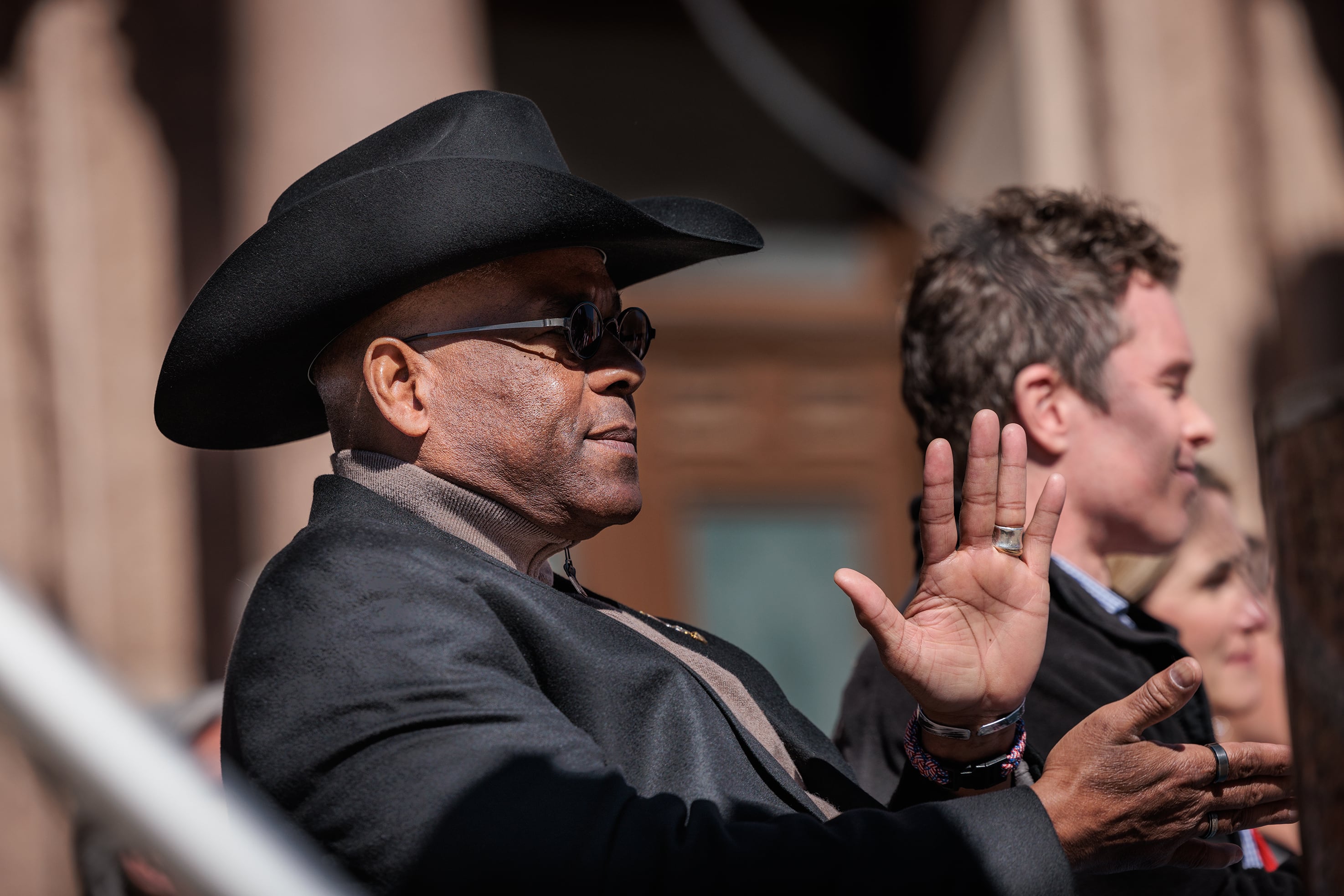 A photograph of a Black man wearing a black hat and suit sitting outside and clapping.