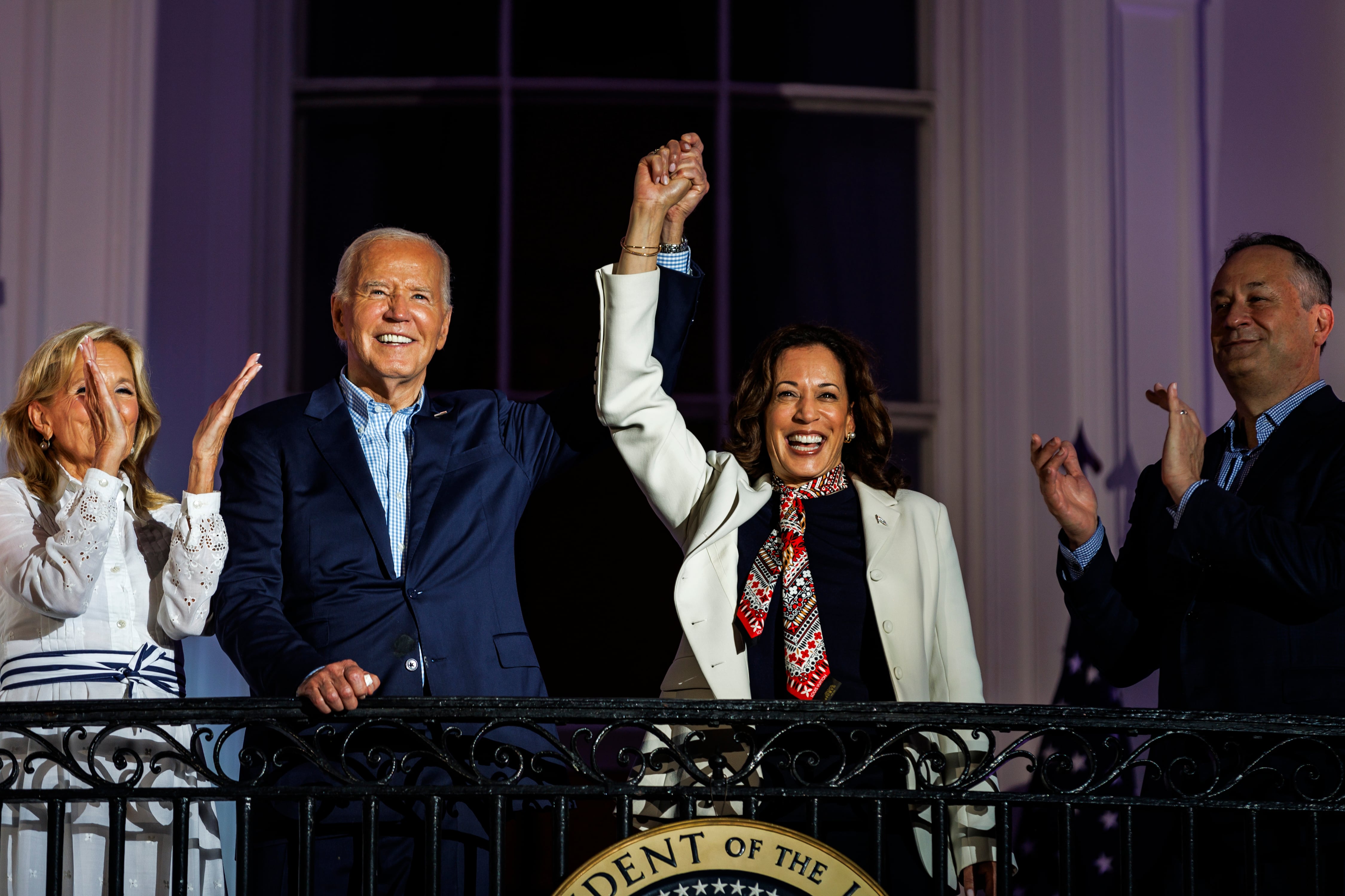 Joe Biden stands on a balcony holding the hand of Kamala Harris high over their heads as Jill Biden and Doug Emhoff stand at their sides