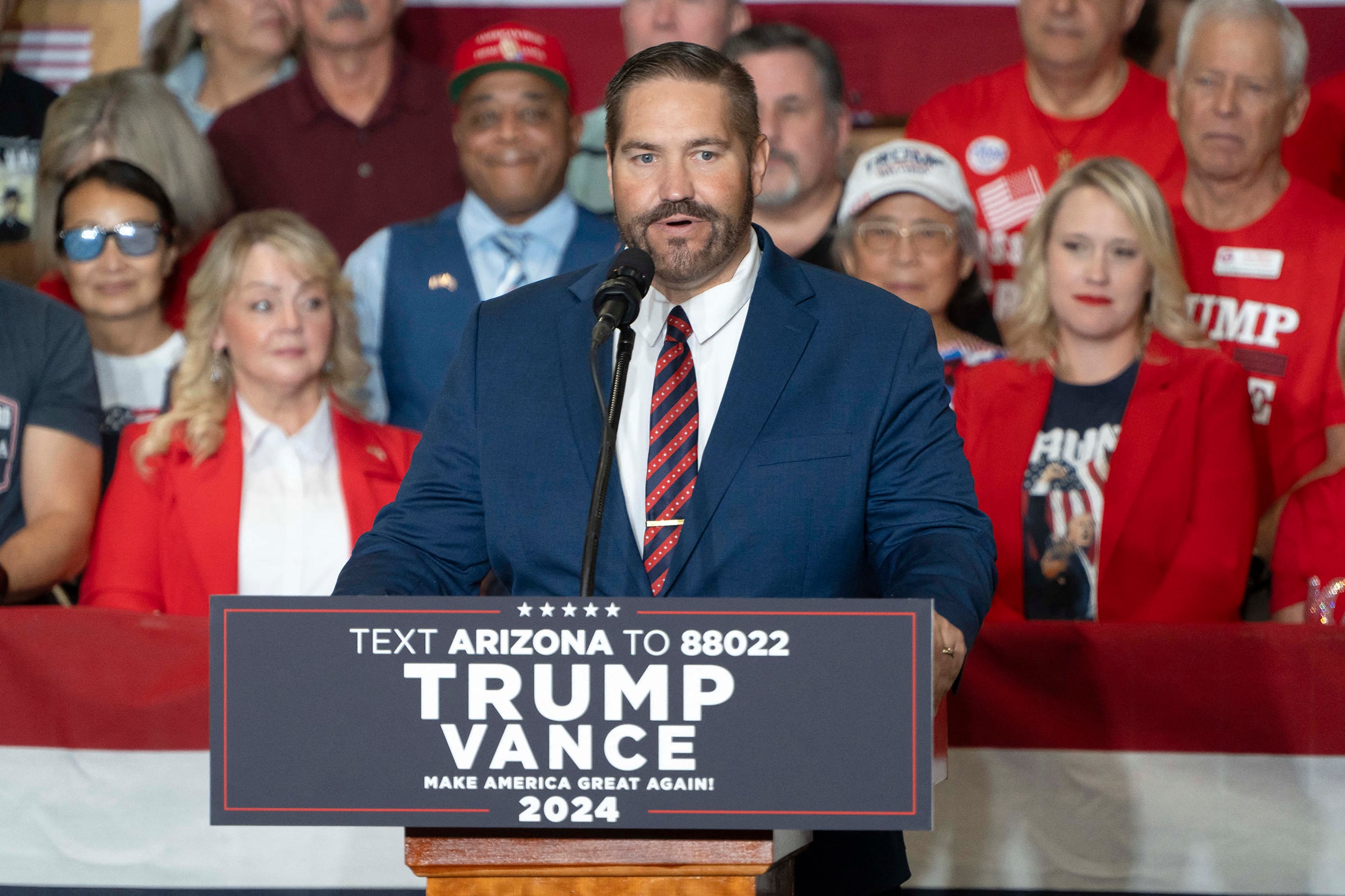 A photograph of a white man in a blue suit stands behind a podium with a large crowd of people sitting in the background.