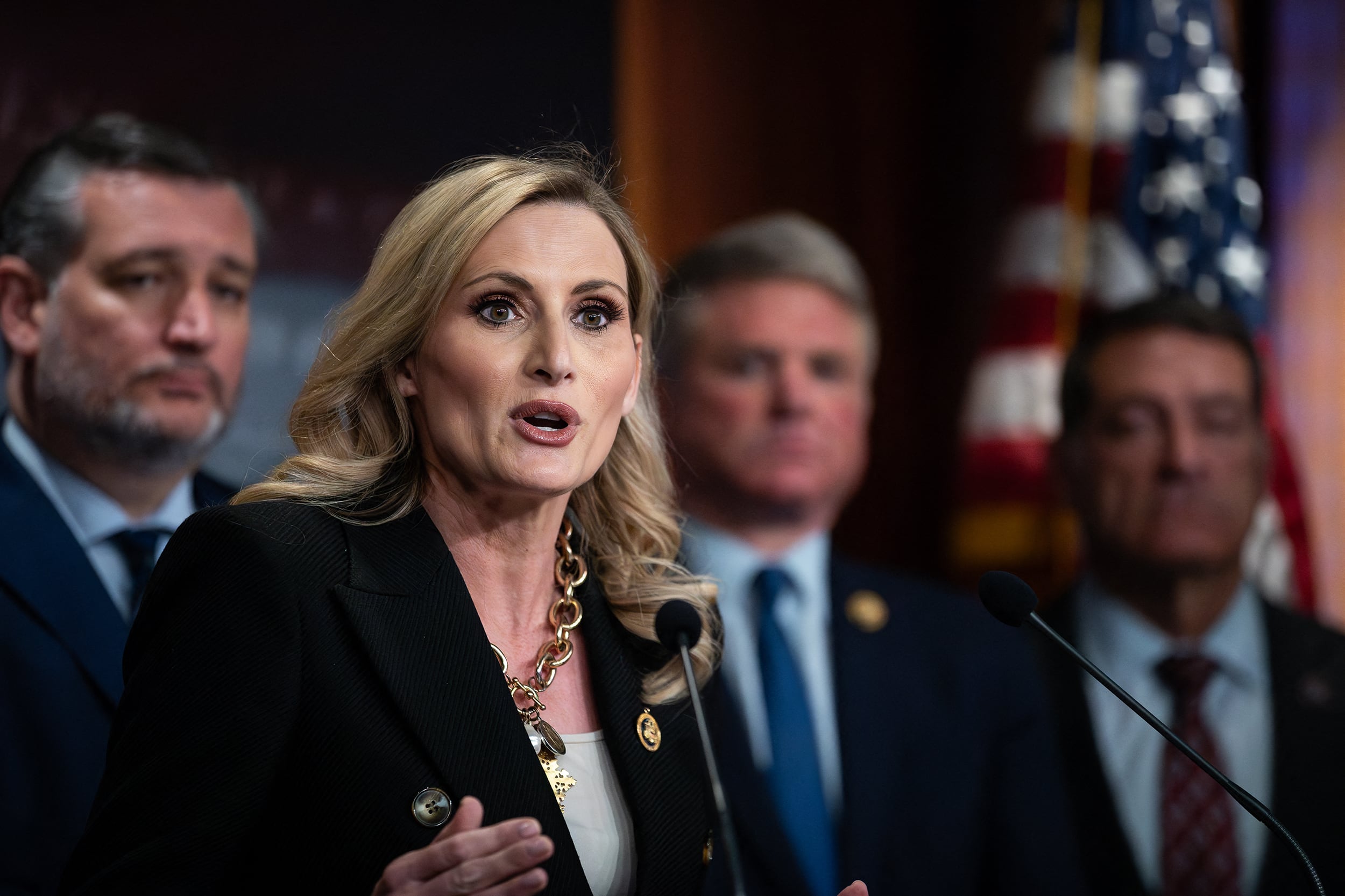 A photograph of a white woman with blonde hair and wearing a dark suit jacket speaks from a microphone with three men in suits standing in the background.
