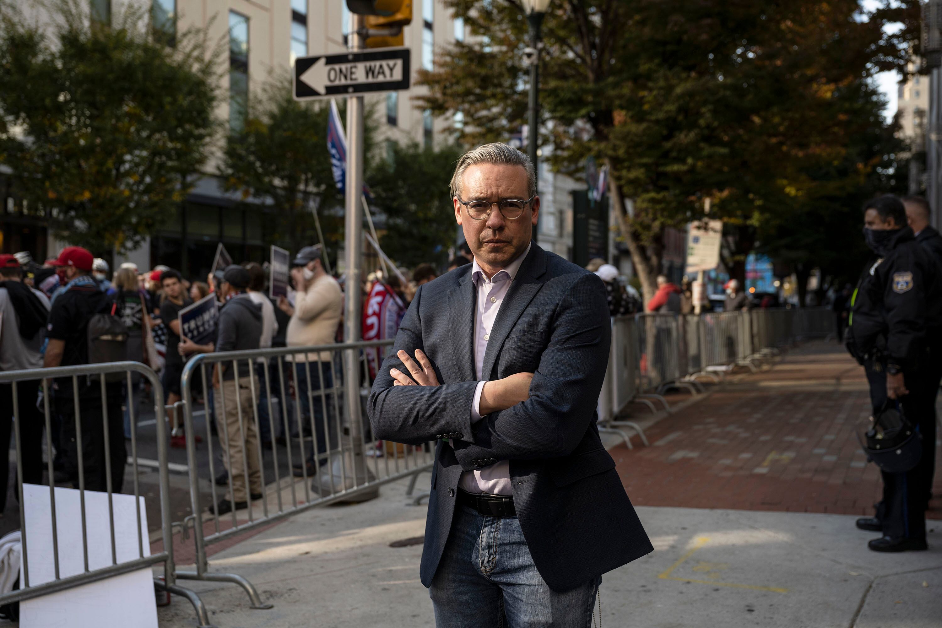 A man wearing a dark suit jacket and glasses poses for a portrait outside of a building in front of a group of people behind a fence.
