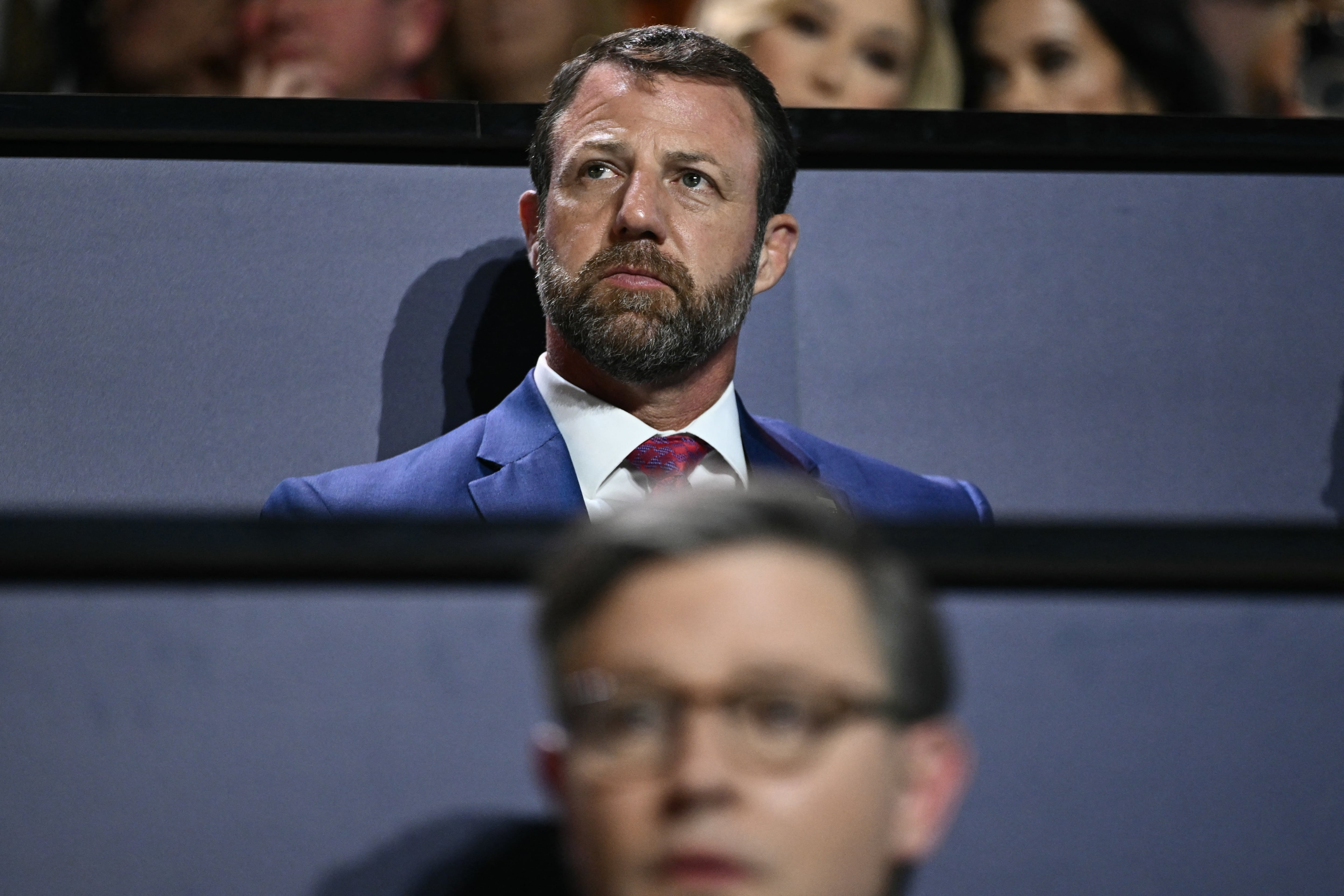 A man in a dark suit jacket sits in a row with another man with glasses out of focus in the foreground.