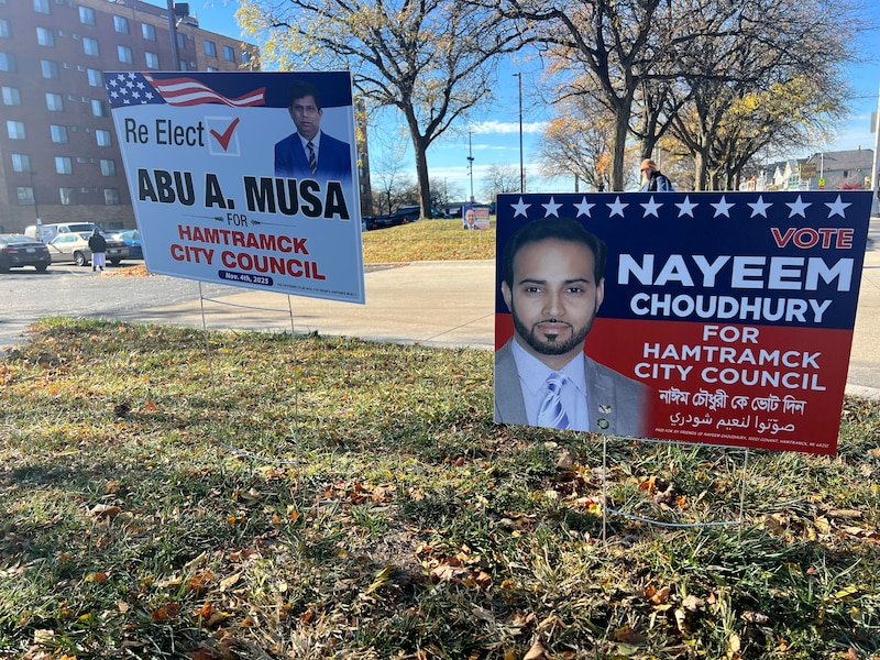 A photograph of two political signs in a grassy lawn outside of a large building in the background.
