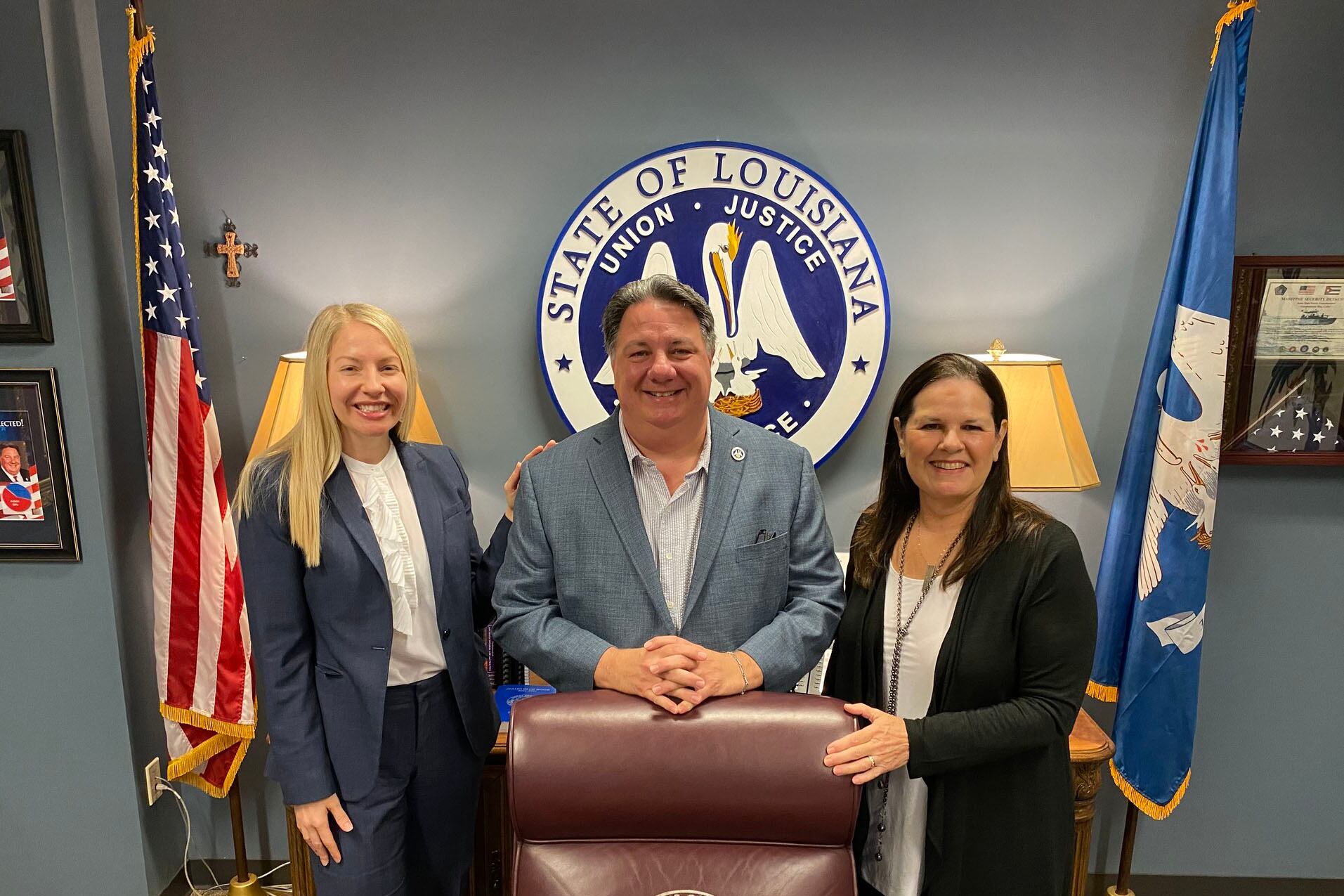 Three people stand side by side behind a podium smiling for a photo with the state of Louisiana seal in the background.