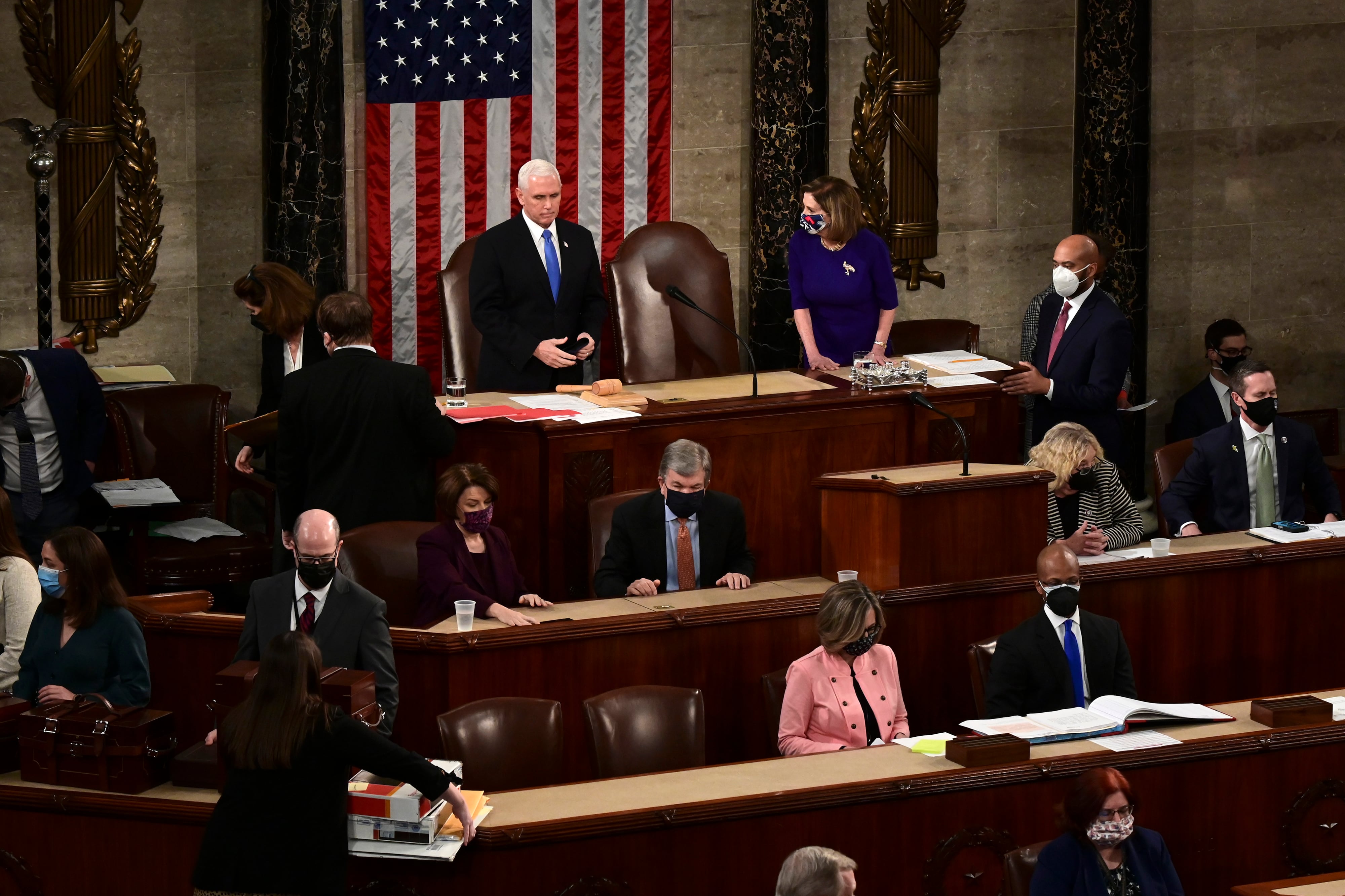 A group of people, some standing and some sitting in a large room with wooden desks stacked on different levels with a large American flag hanging in the background.