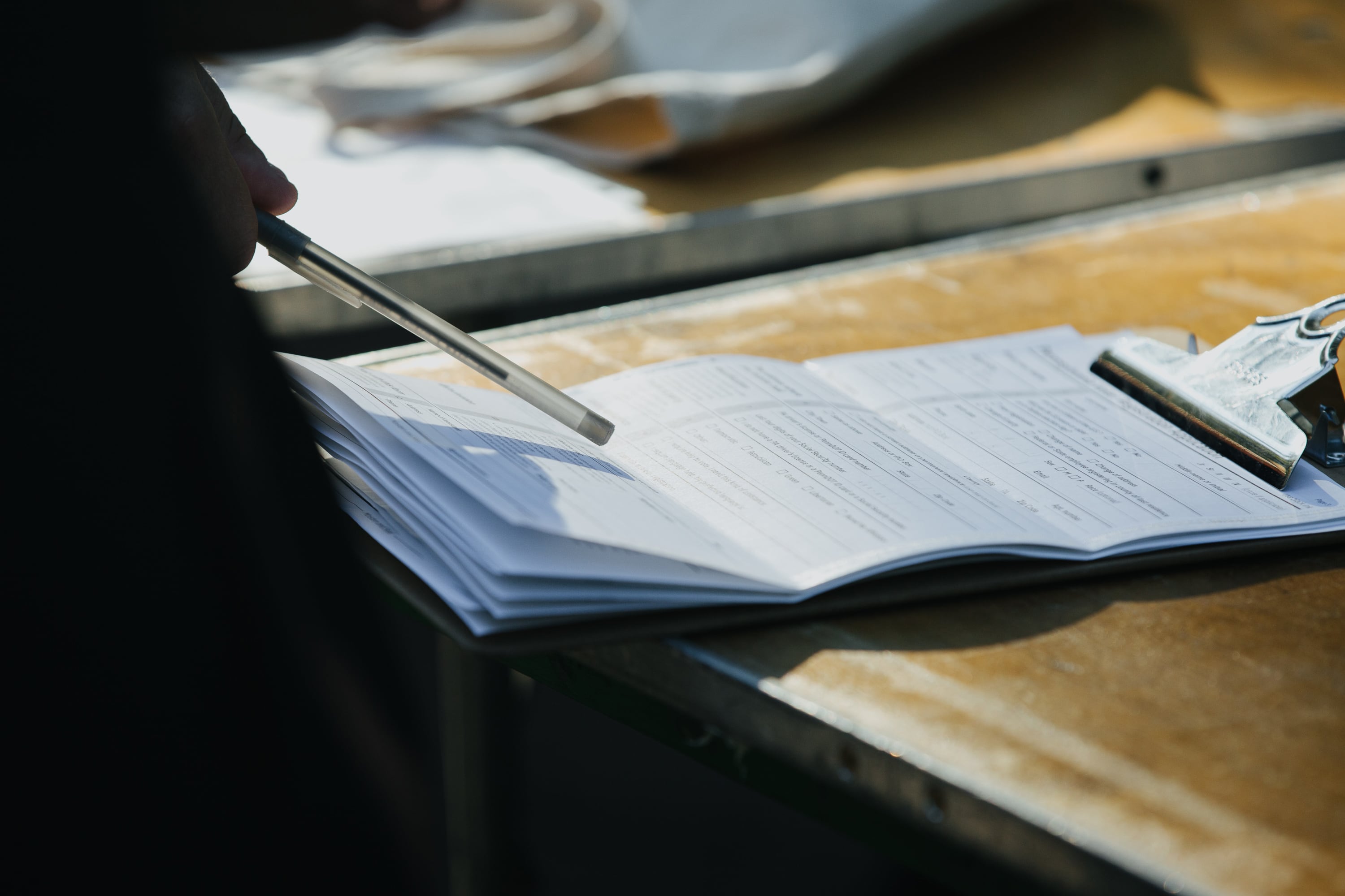 A hand holds a pen over a clipboard with white pieces of paper.