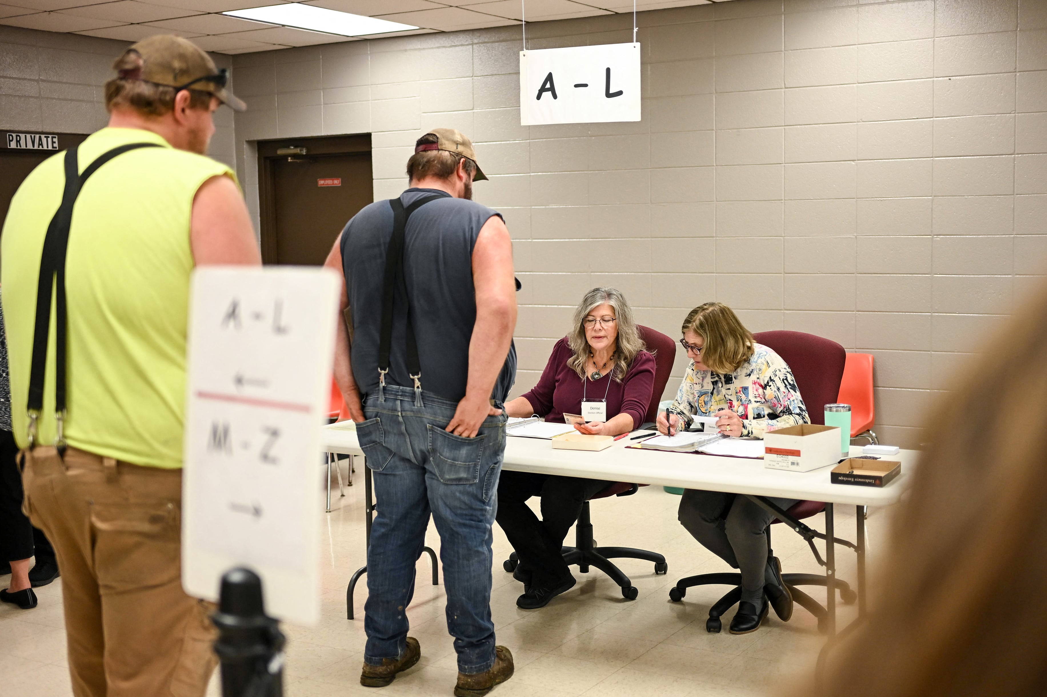 Two men wearing suspenders stand in line in front of two white women who are sitting at a table helping voters vote.