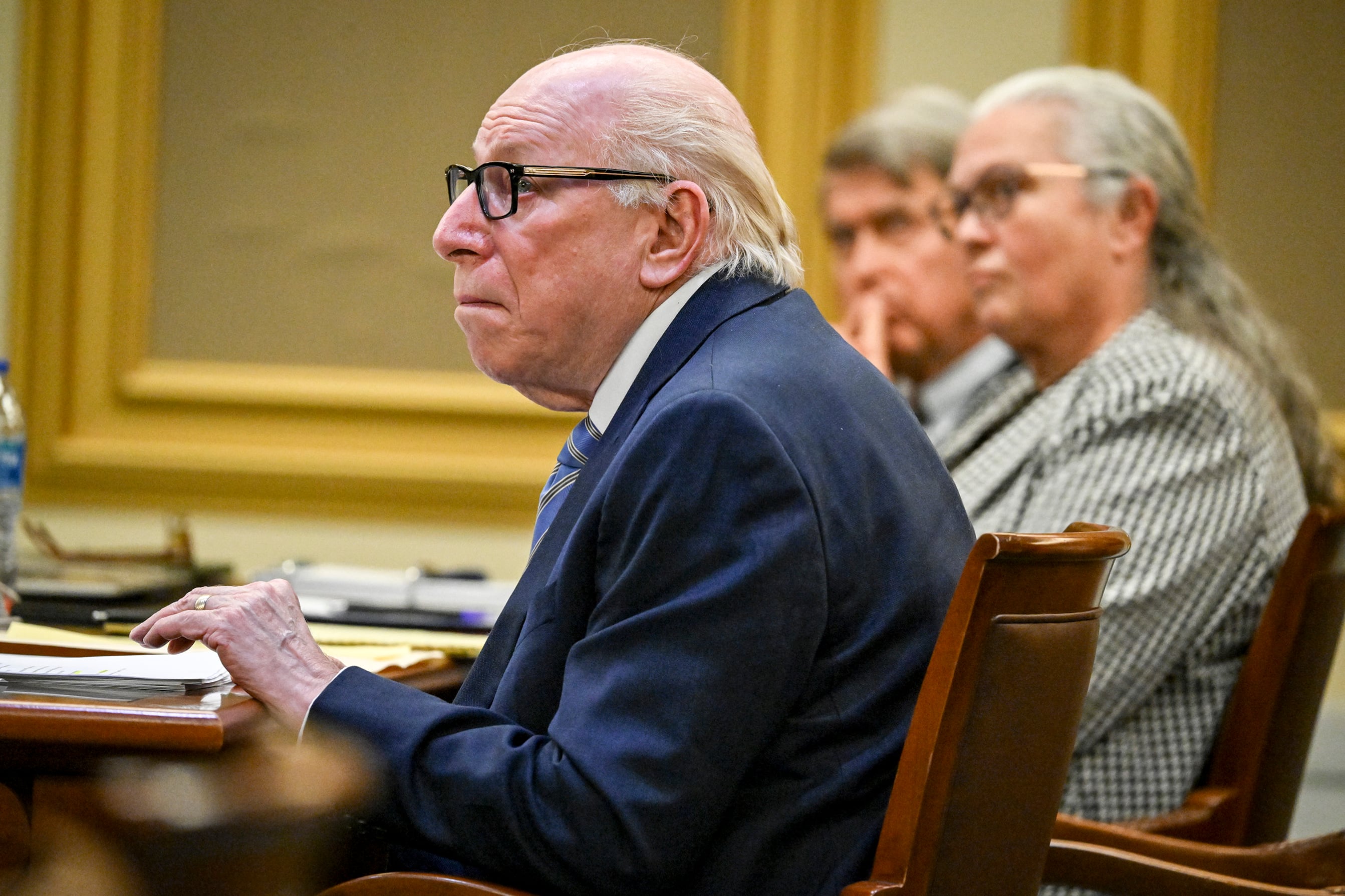 A white man in a suit sits at a wooden table in a court room.