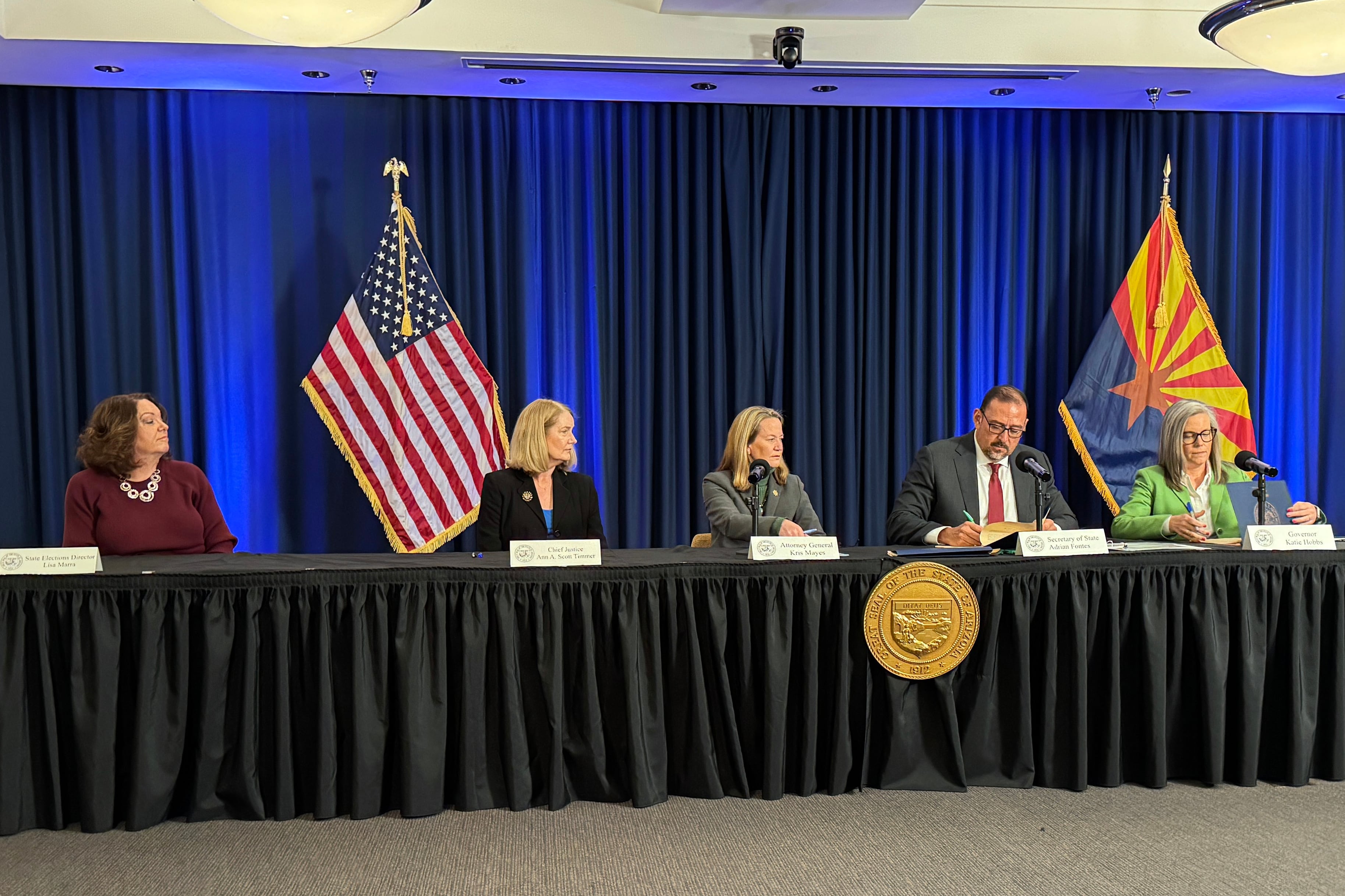 Five adults in business attire sit at a long table in front of a blue backdrop with an American and Arizona flag.