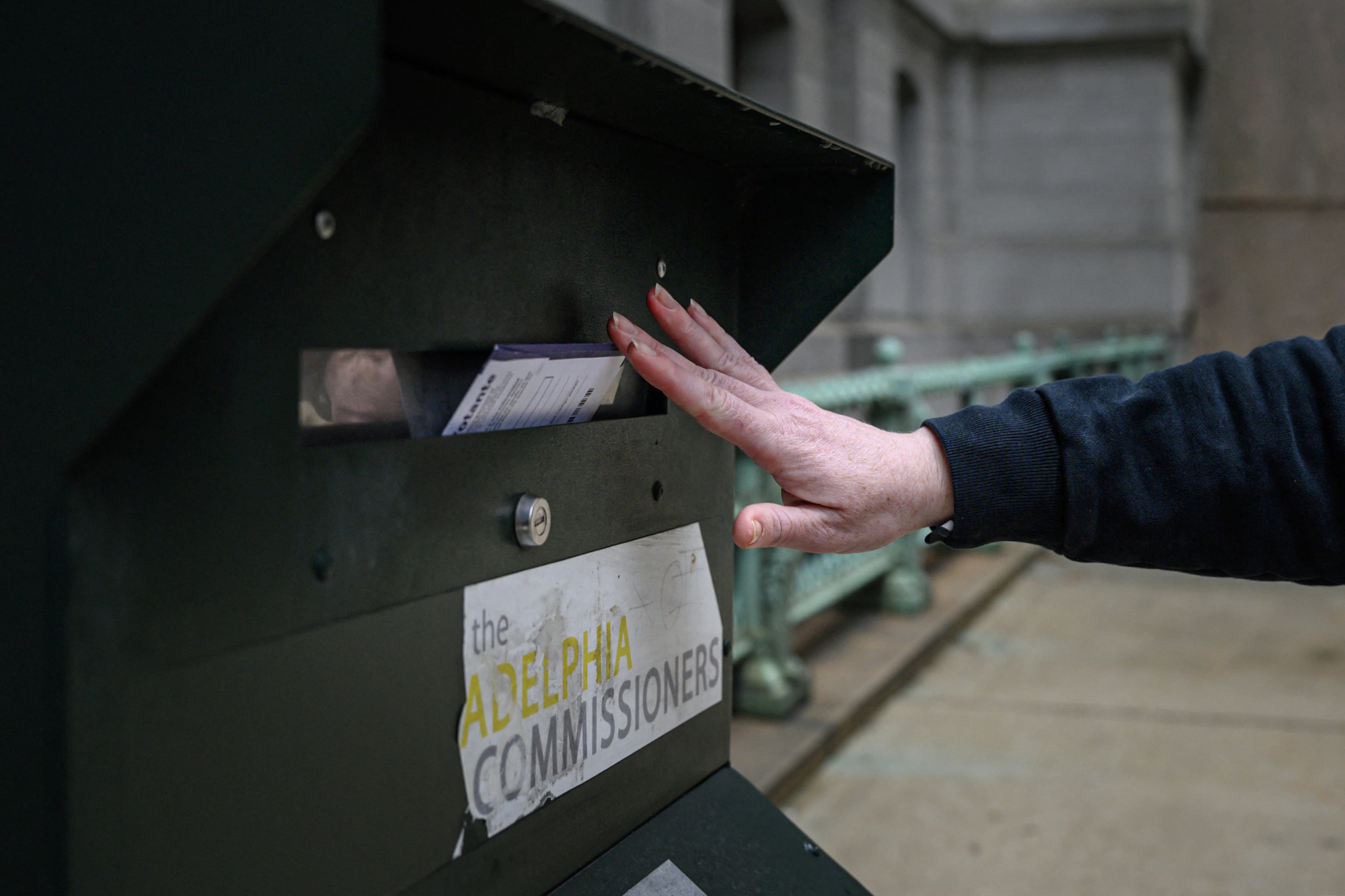 An up-close photo of a hand pushing a white mail-in ballot into a secure mail ballot drop box outside a large stone building.