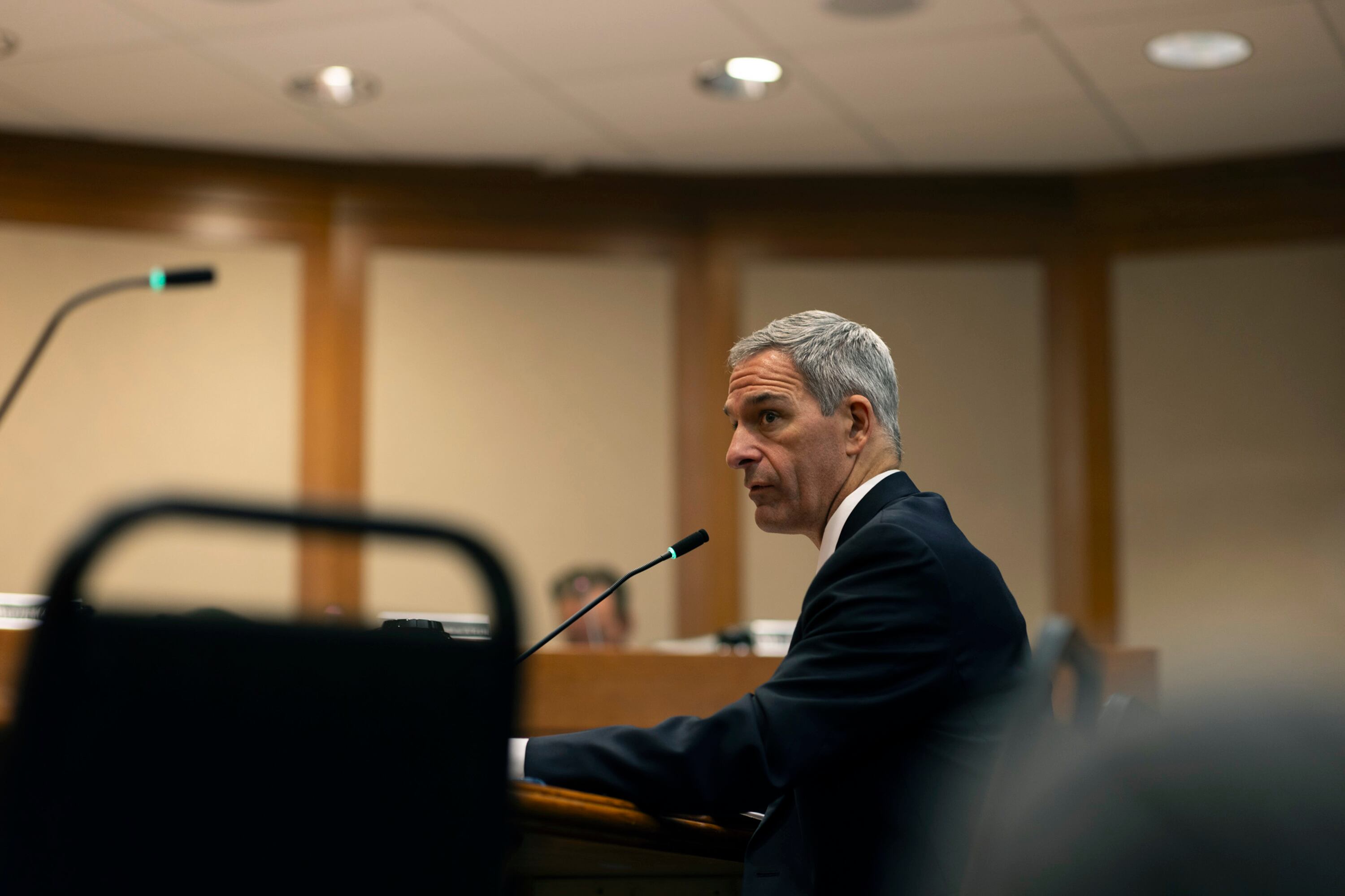 A man with gray hair seated at a table in a meeting room listens to an unseen speaker