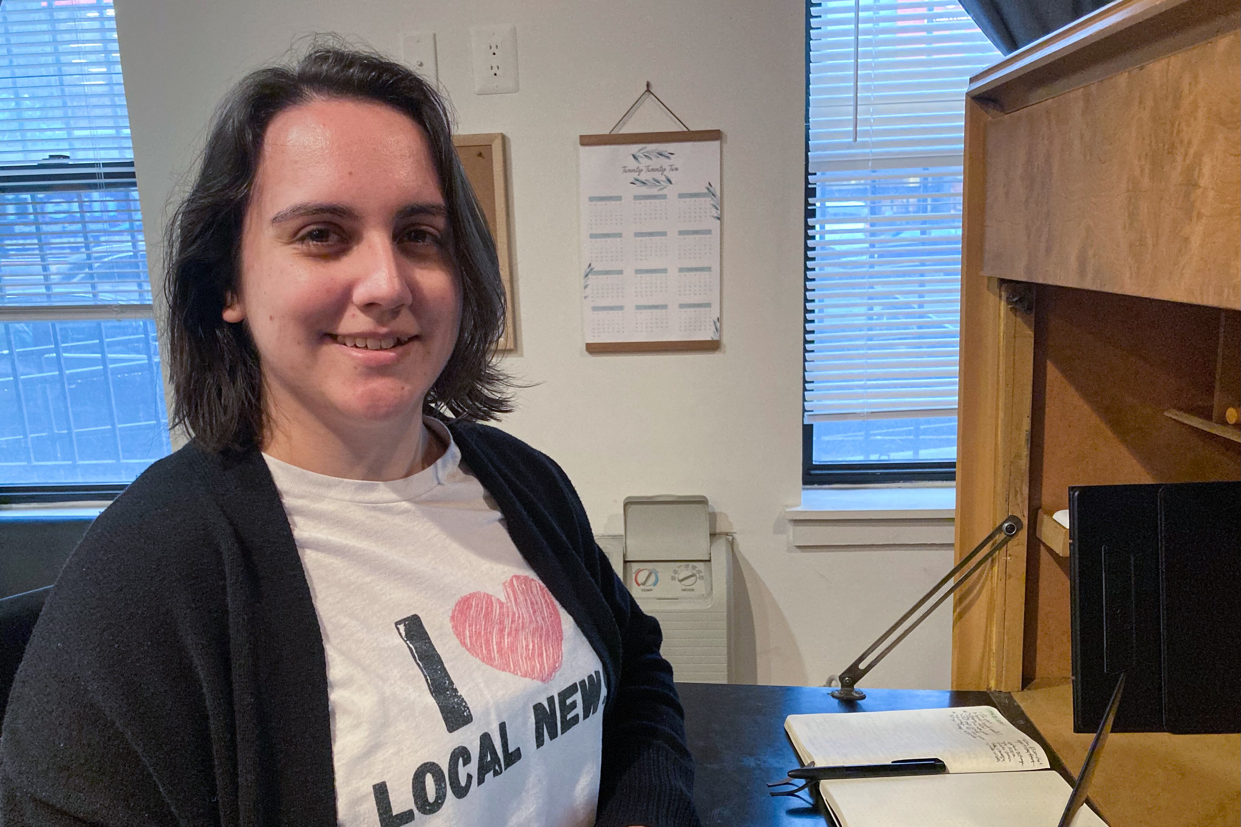 Woman sitting at a desk wearing a T-shirt that reads “I love local news”