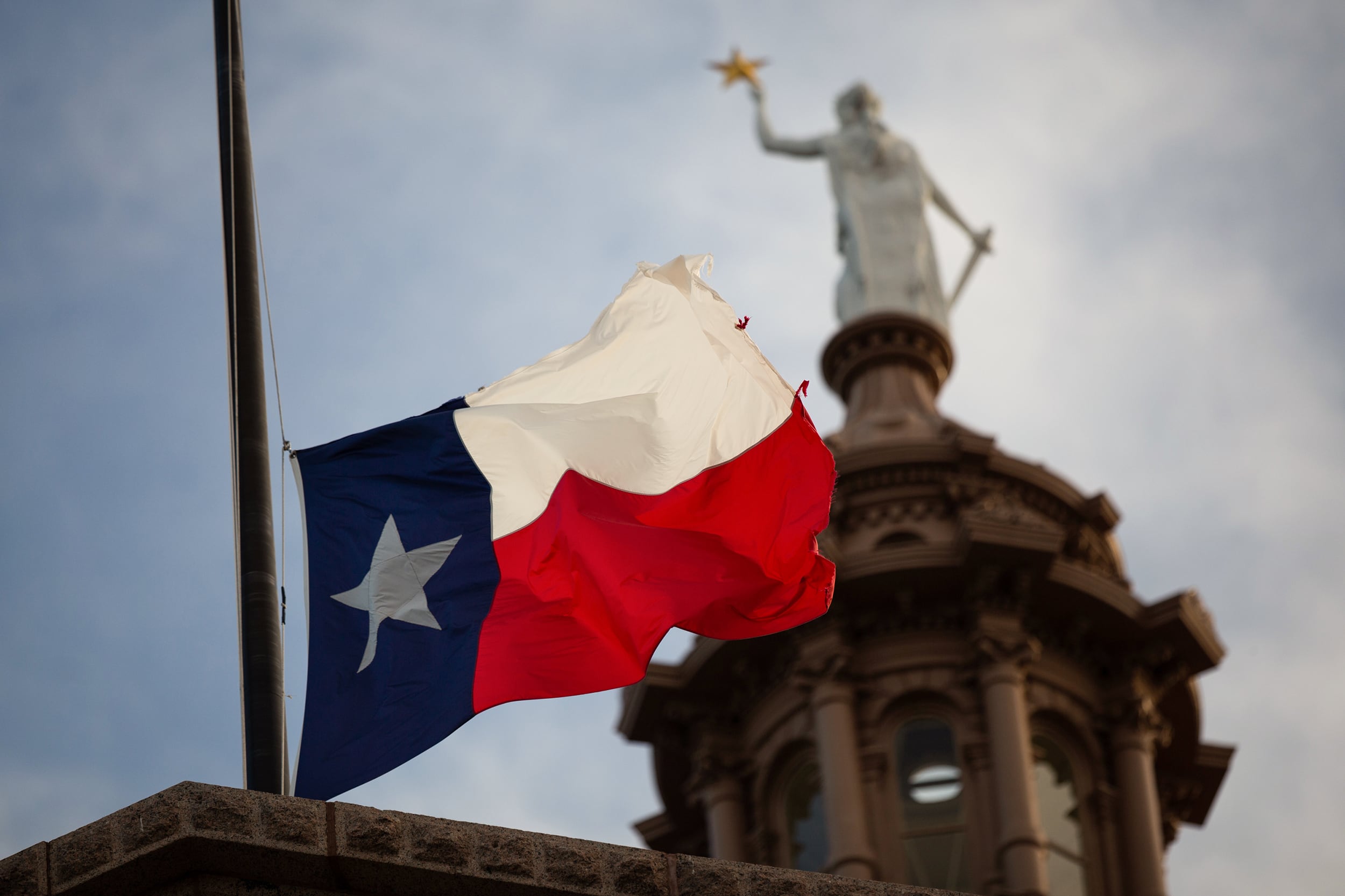A photograph of the top of the Texas State Capitol statue with the Texas state flag in focus in the foreground.