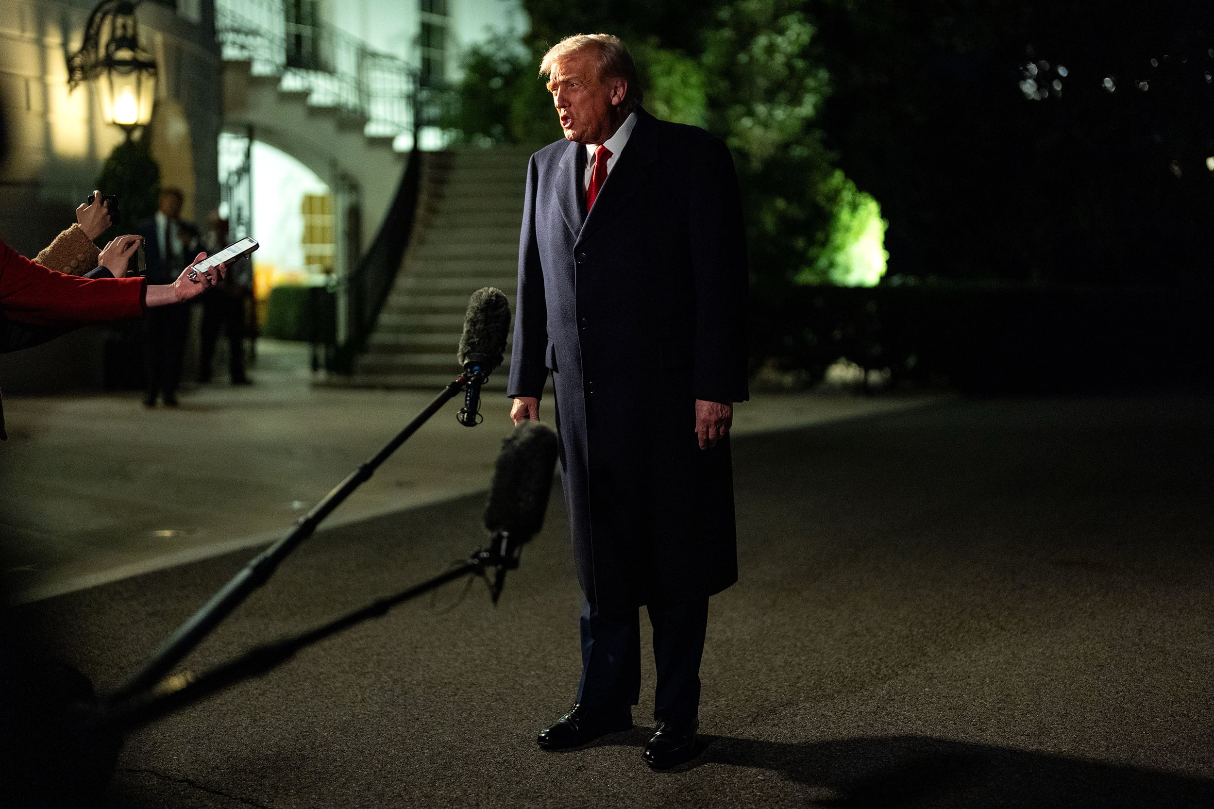 A photograph of a white man in a suit and long jacket standing outside in the dark with the White House in the background and reporters on the left.