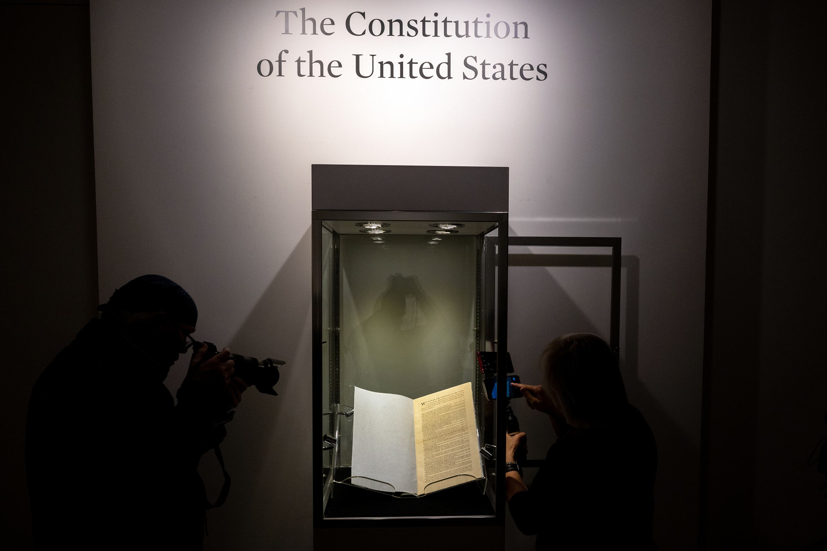 Two people stand next to a glass case with the first paper copy of the US constitution with the words on the wall above that reads "The Constitution of the United States."