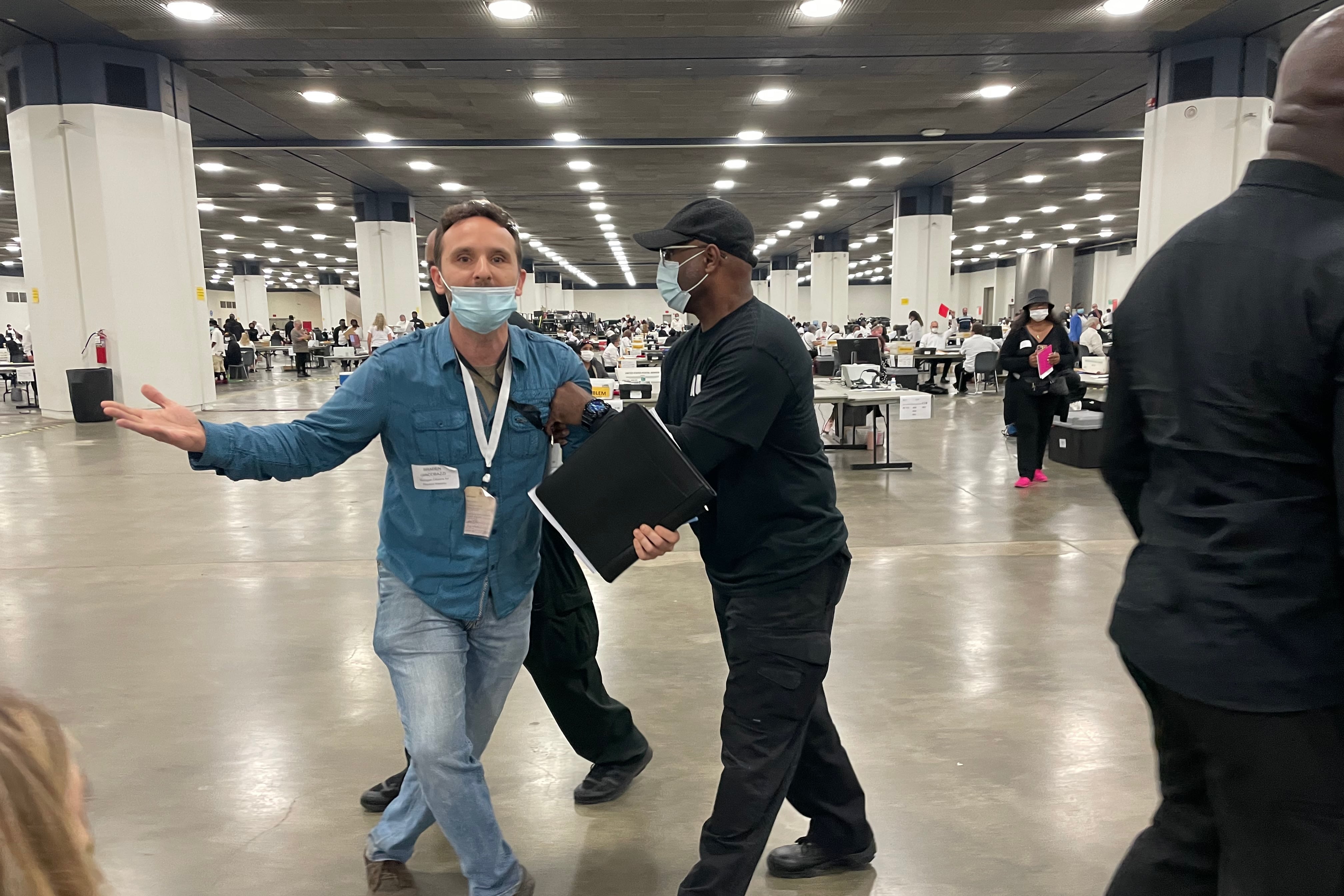 A man in a blue shirt and jeans gestures with his arms up while a man ushers him away.