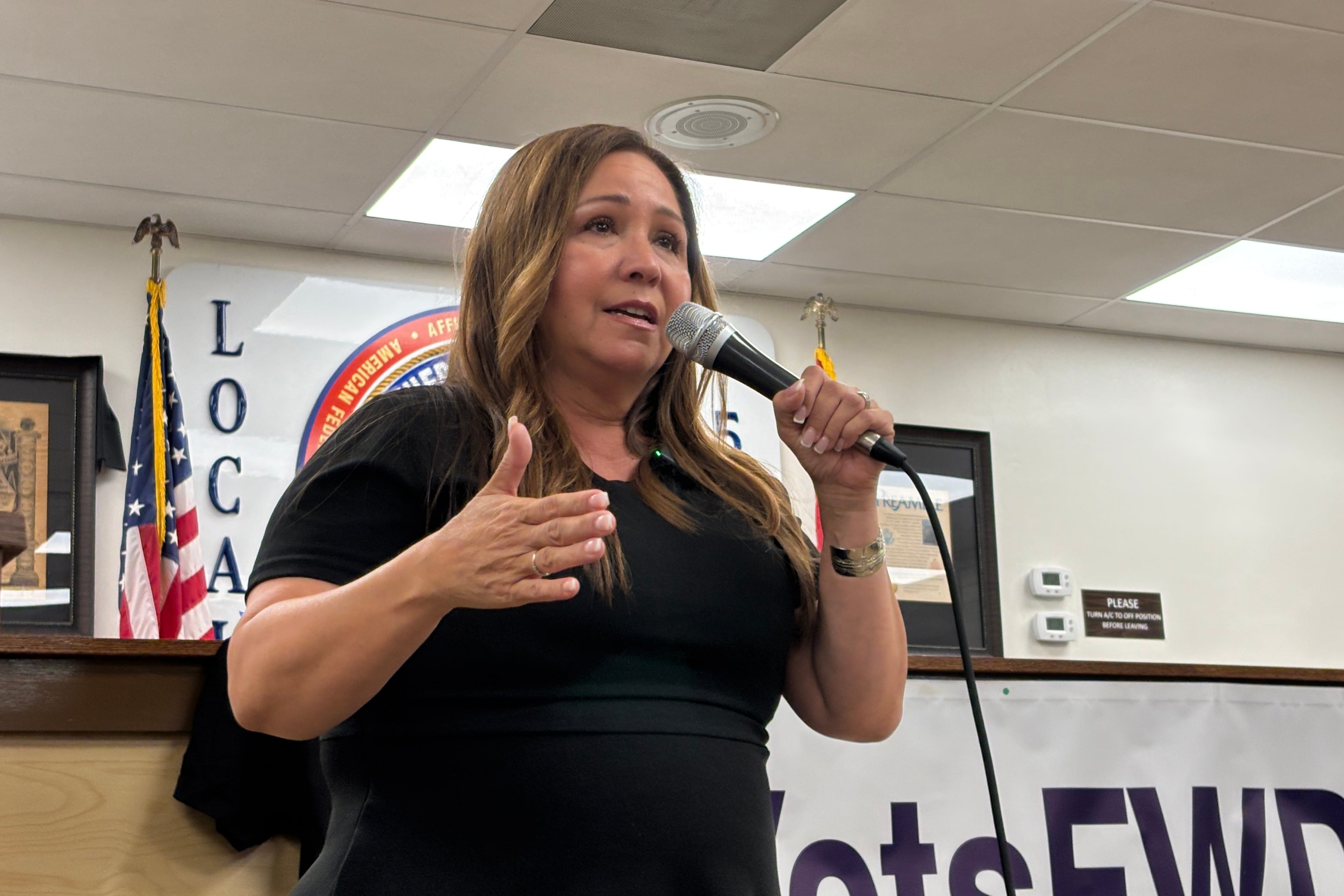 A photograph of a woman speaking into a microphone at the front of a room.