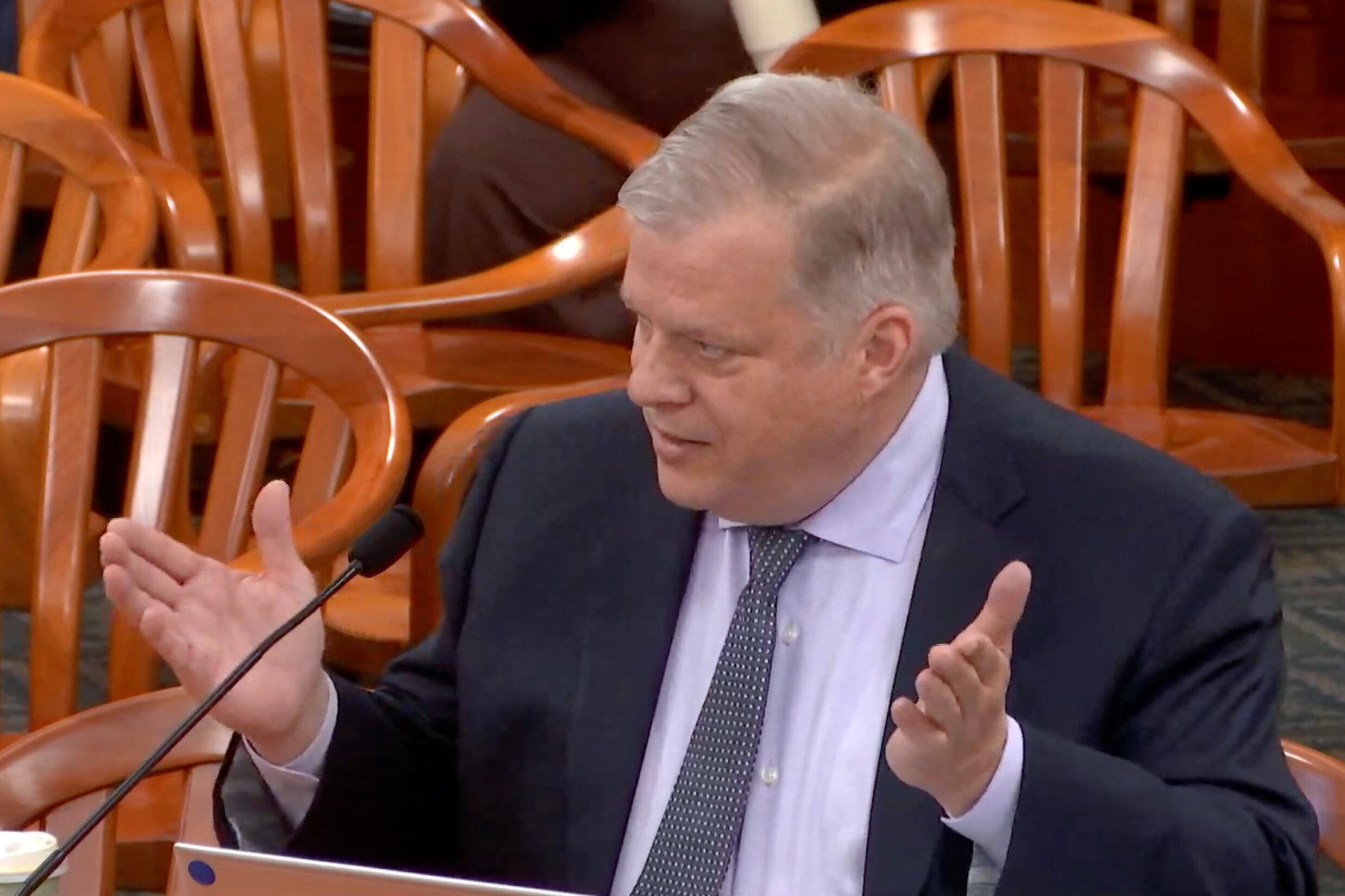 A white man wearing a dark suit and tie speaks from a podium with his hands spread out.