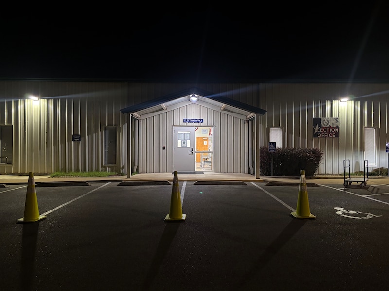 A photograph of the front entrance to a building at night with two lights on each side of the entrance and yellow safety cones in a line in the foreground.