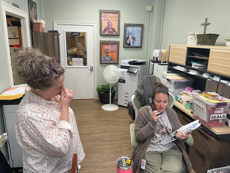 A photograph of two white women working in an office together. One person is on the phone and holding a large stack of papers and one is holding her head in a look of exhaustion.