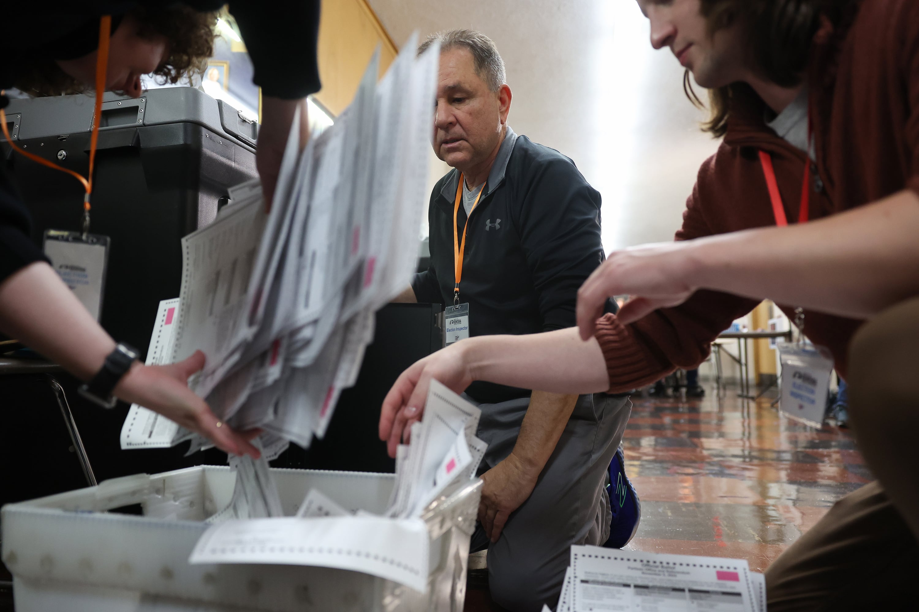 A man in a gray sweat jacket watches as people work with ballots.