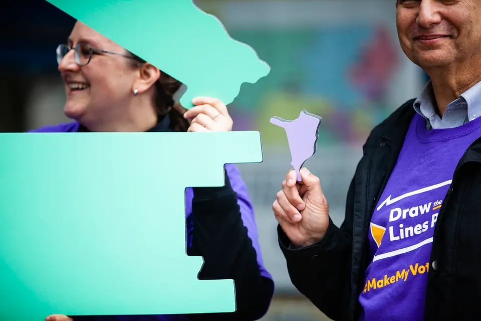 A man and woman wearing purple T-shirts that read “Draw the Lines” hold large green or purple puzzle pieces shaped like Pennsylvania congressional districts.