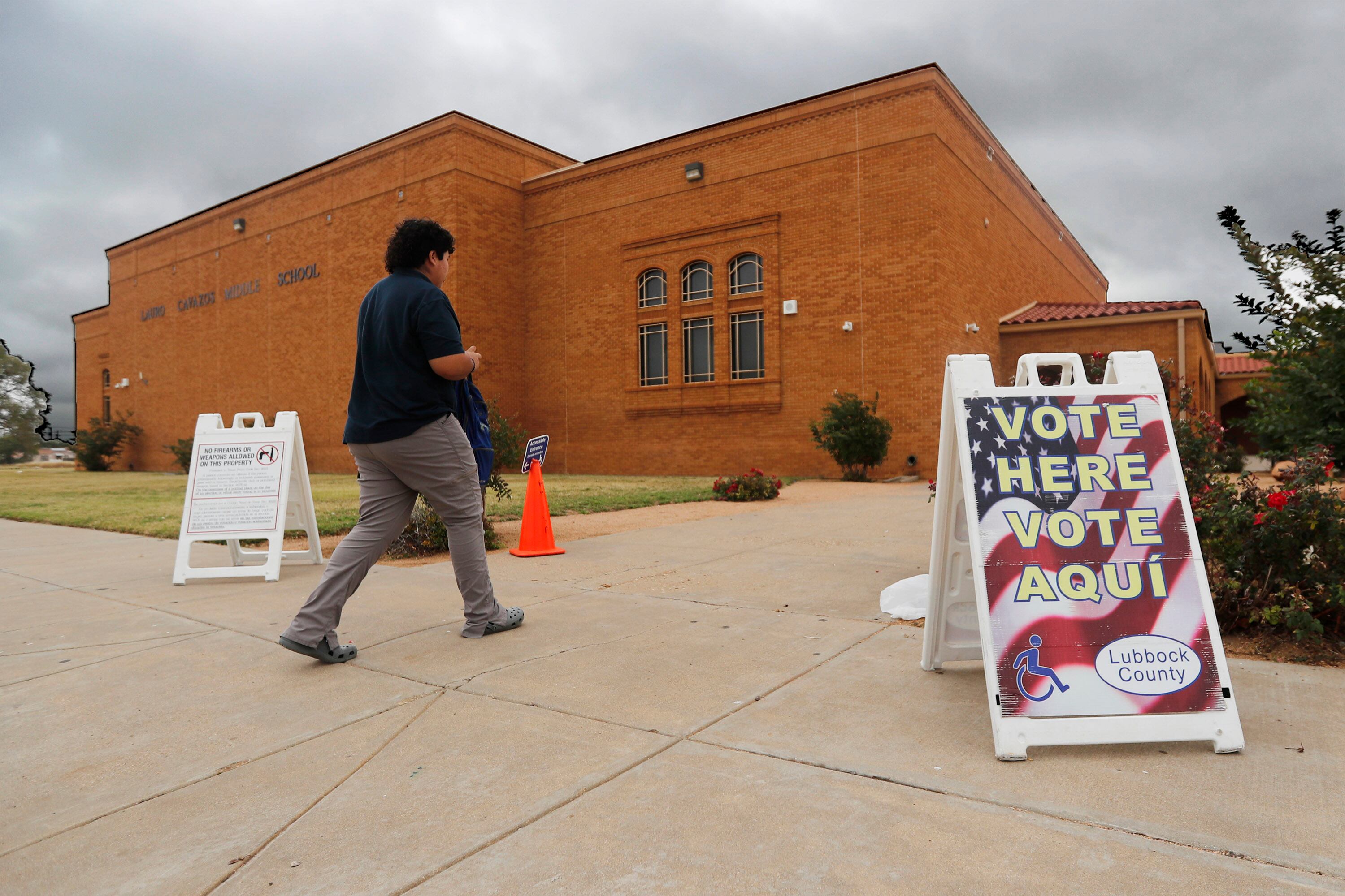 A person wearing a dark shirt and pants walks outside behind a "vote here vote aqui" sign and in front of a red brick building.